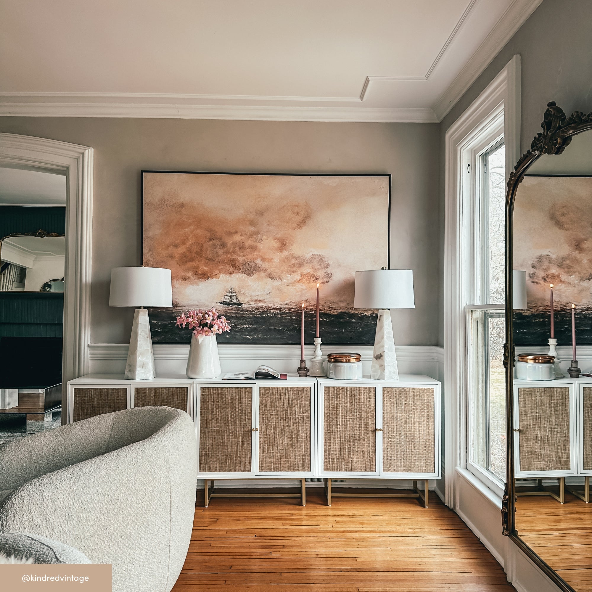 A stylish living room with a white sideboard, two table lamps, pink flowers, and candles, beneath a large painting. A curved armchair sits nearby and a tall mirror reflects a window with natural light.