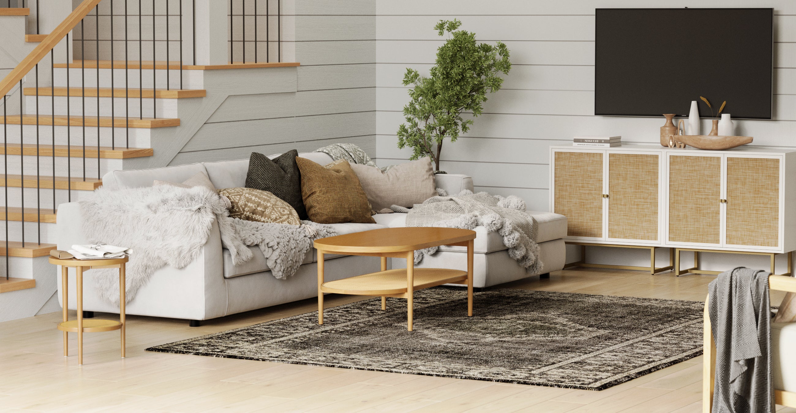 A cozy living room with a sectional sofa covered in neutral pillows and throws, a wooden coffee table, side table, TV on the wall, a cabinet, indoor plant, and a patterned rug on light wood floors near a staircase.