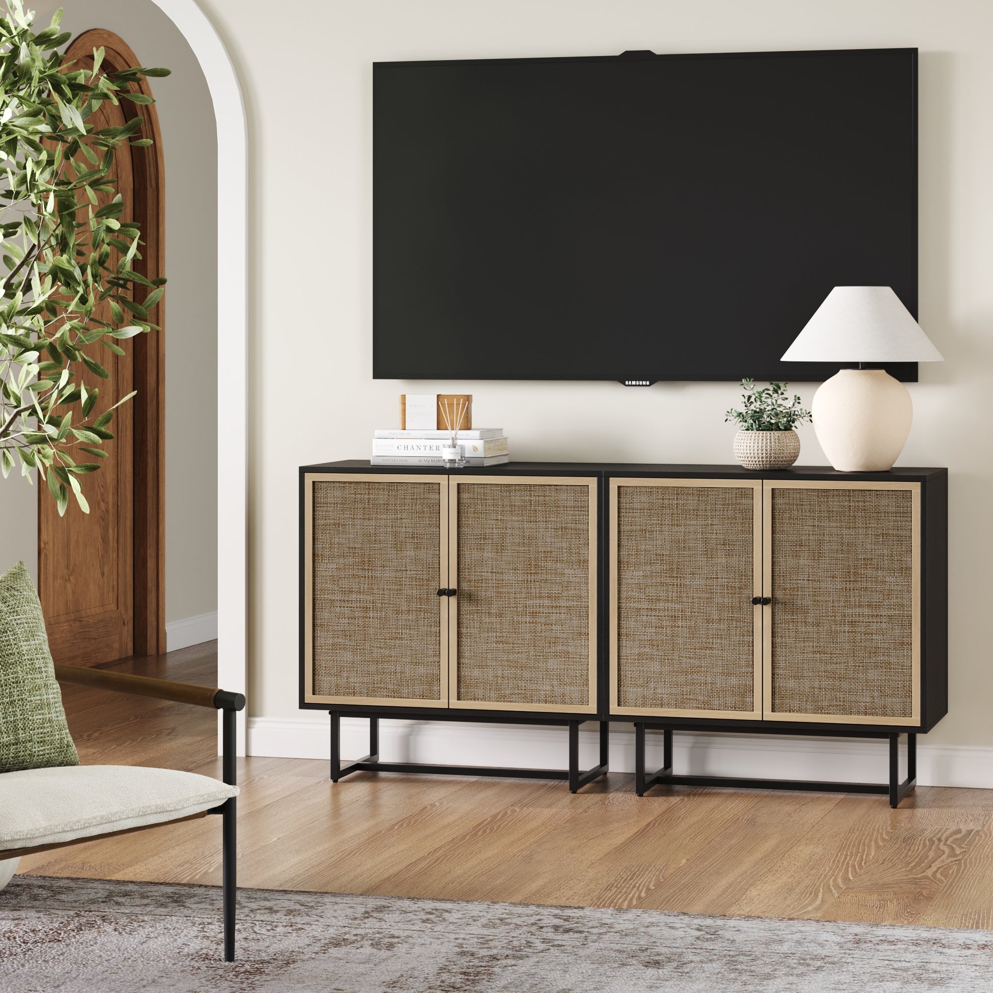 A modern living room showcases a wall-mounted TV above the Nathan James Rattan TV Console Credenza Black (Set of 2), styled with books, a vase, and a lamp. A green plant and chair are on the left, with an arched doorway in the background.