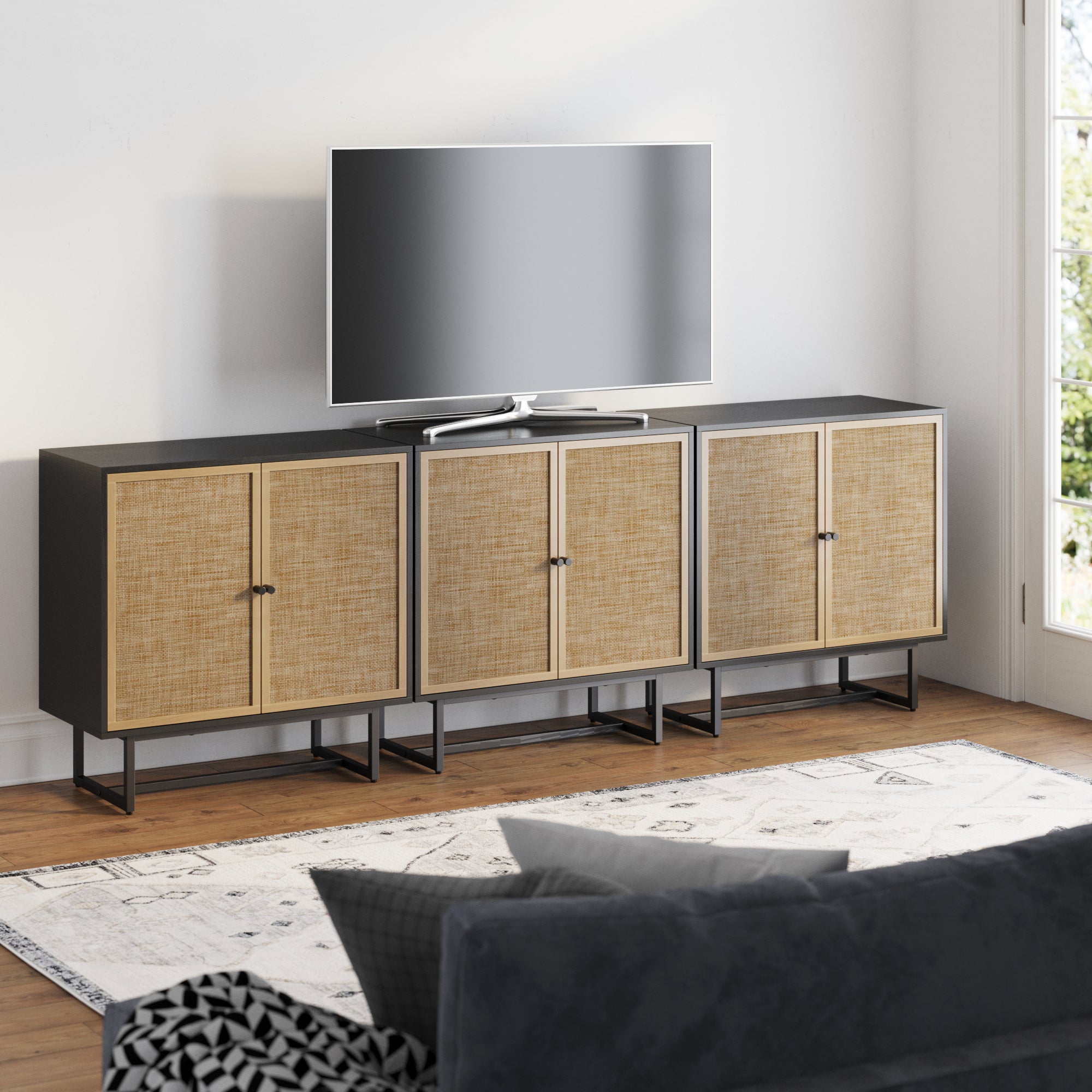 A modern living room features the Nathan James Rattan TV Console Credenza Black (Set of 3), with four woven rattan cabinet doors that blend bohemian style and sleek design. A flat-screen TV, gray sofa, and patterned rug complete the look on a wooden floor.