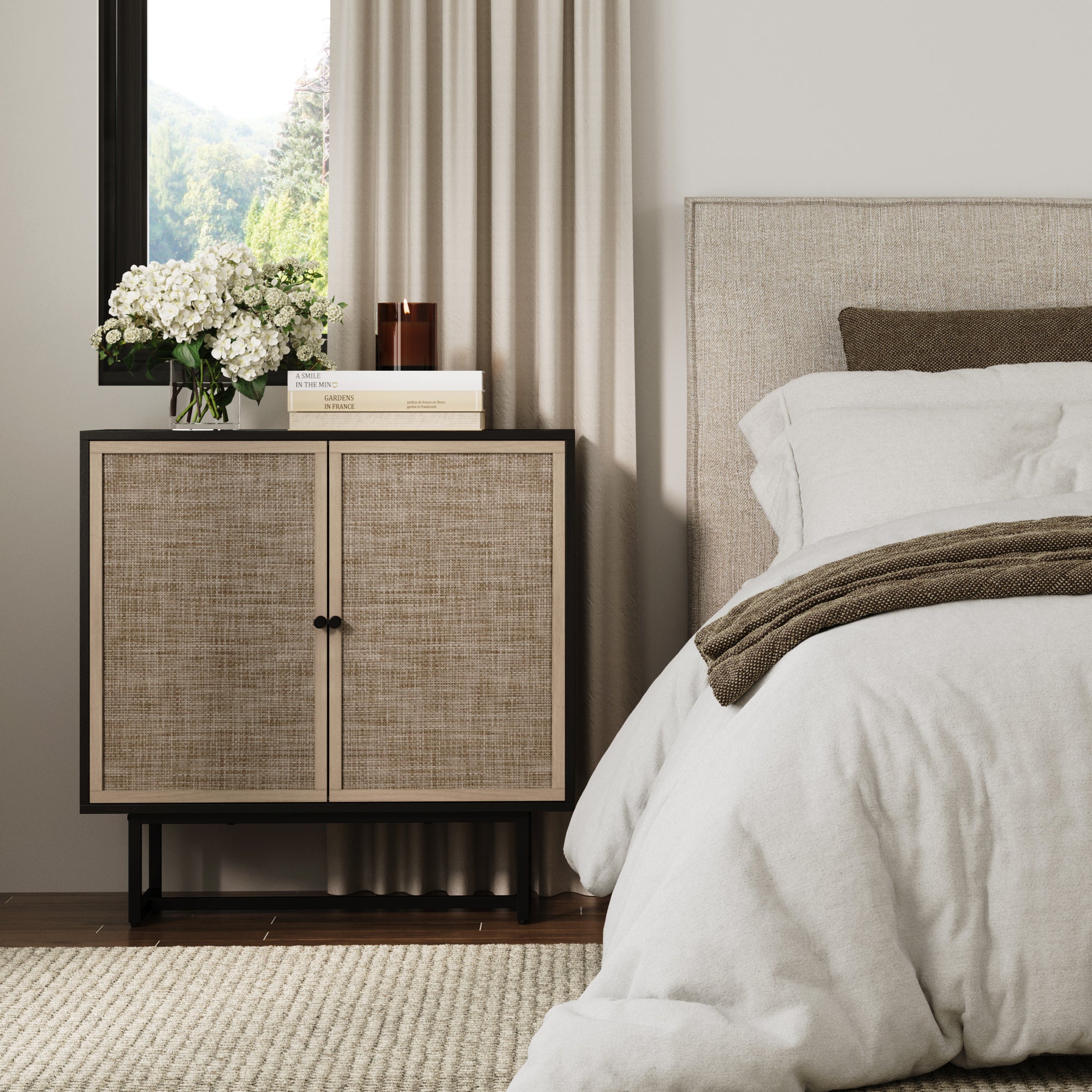 A cozy bedroom with a bed dressed in white and beige bedding, a woven-textured nightstand with flowers, books, and a candle on top, beige curtains, and a window with a view of greenery outside.
