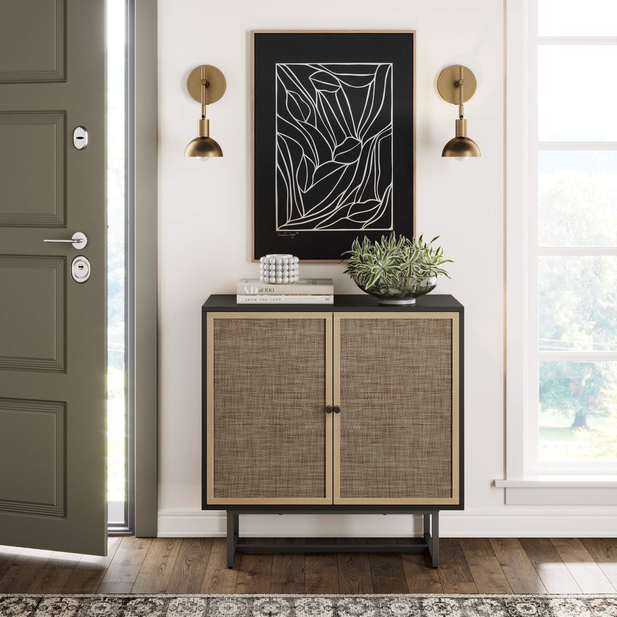 A modern entryway features an olive door, the Nathan James Wood & Rattan Storage Cabinet in black, a plant, books, abstract line wall art, two brass sconces, and a window bringing in natural light.