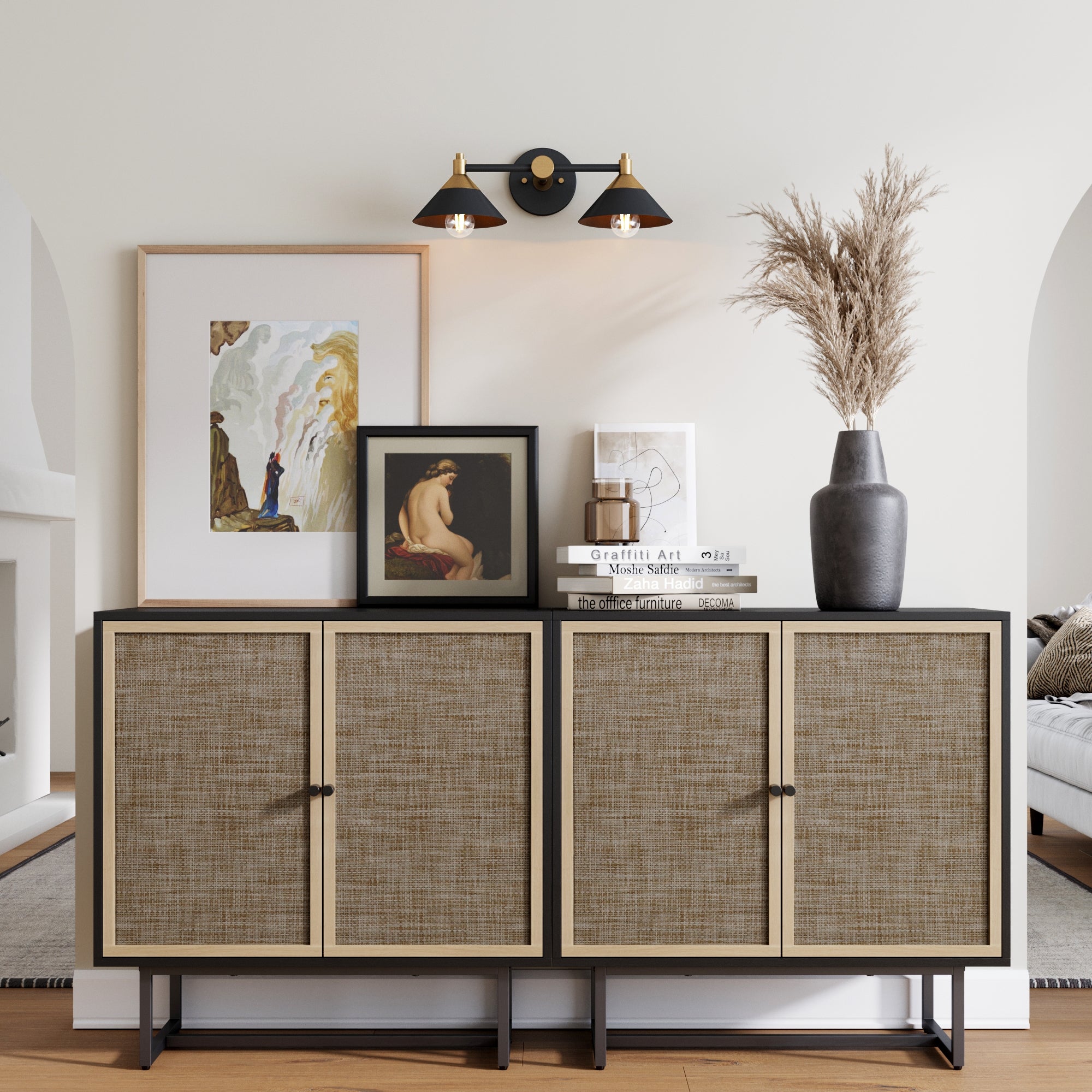 A modern sideboard with woven rattan doors displays framed art, stacked books, and a tall vase with dried pampas grass. Above, a black and gold wall sconce with two lights is mounted on a light-colored wall.