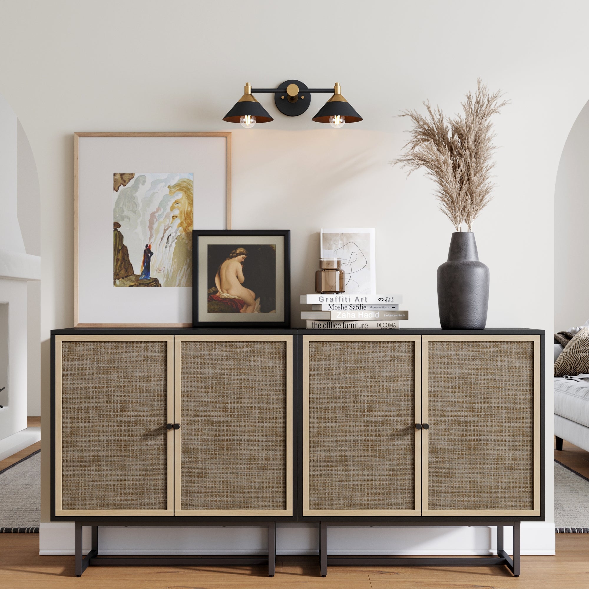 The Nathan James Rattan TV Console Credenza in black (set of 2) features woven doors and is styled with framed art, decorative books, a ceramic vase of dried grasses, and sits against a white wall under a black and gold wall sconce.