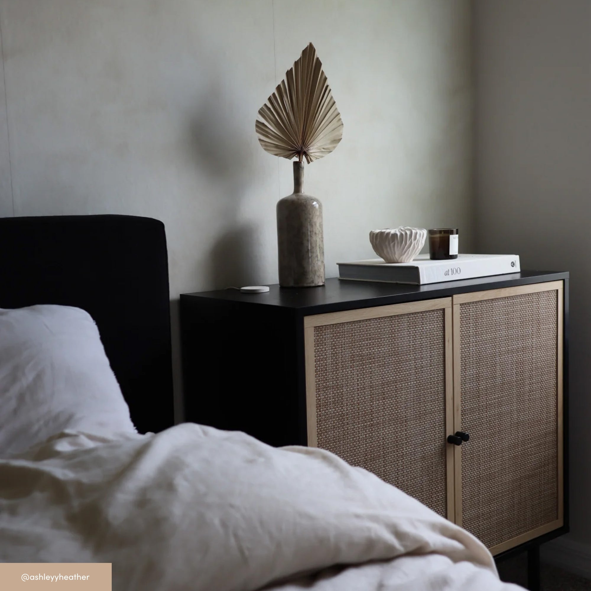 A cozy bedroom scene with a bed in the foreground, white bedding, and a bedside table holding a tall vase with a dried palm leaf, a stacked book, a ceramic bowl, and a small candle.