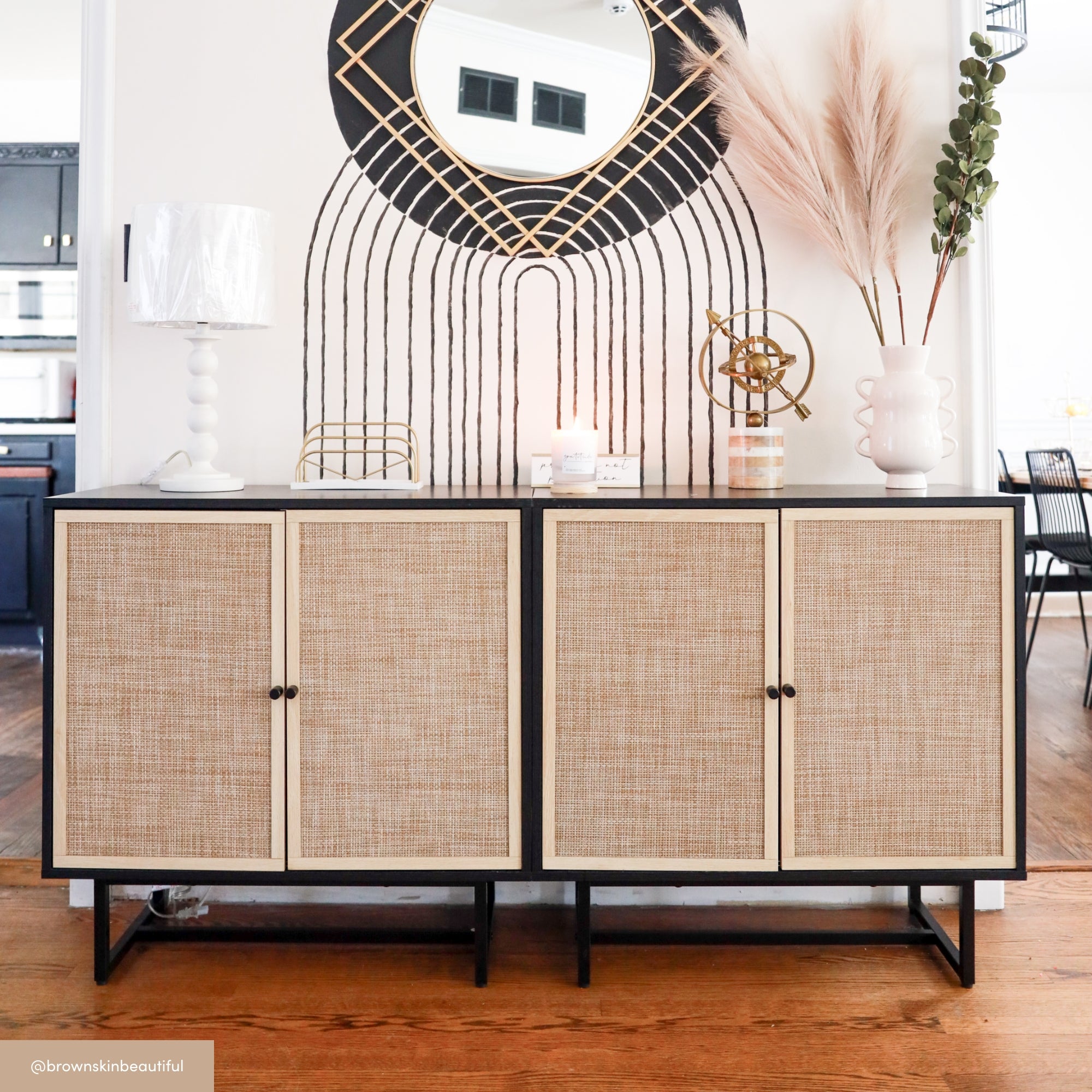 A modern sideboard with wicker doors sits against a white wall, topped with decor items including a lamp, candles, and vases. Above, a round mirror with black and gold geometric designs is flanked by pampas grass.