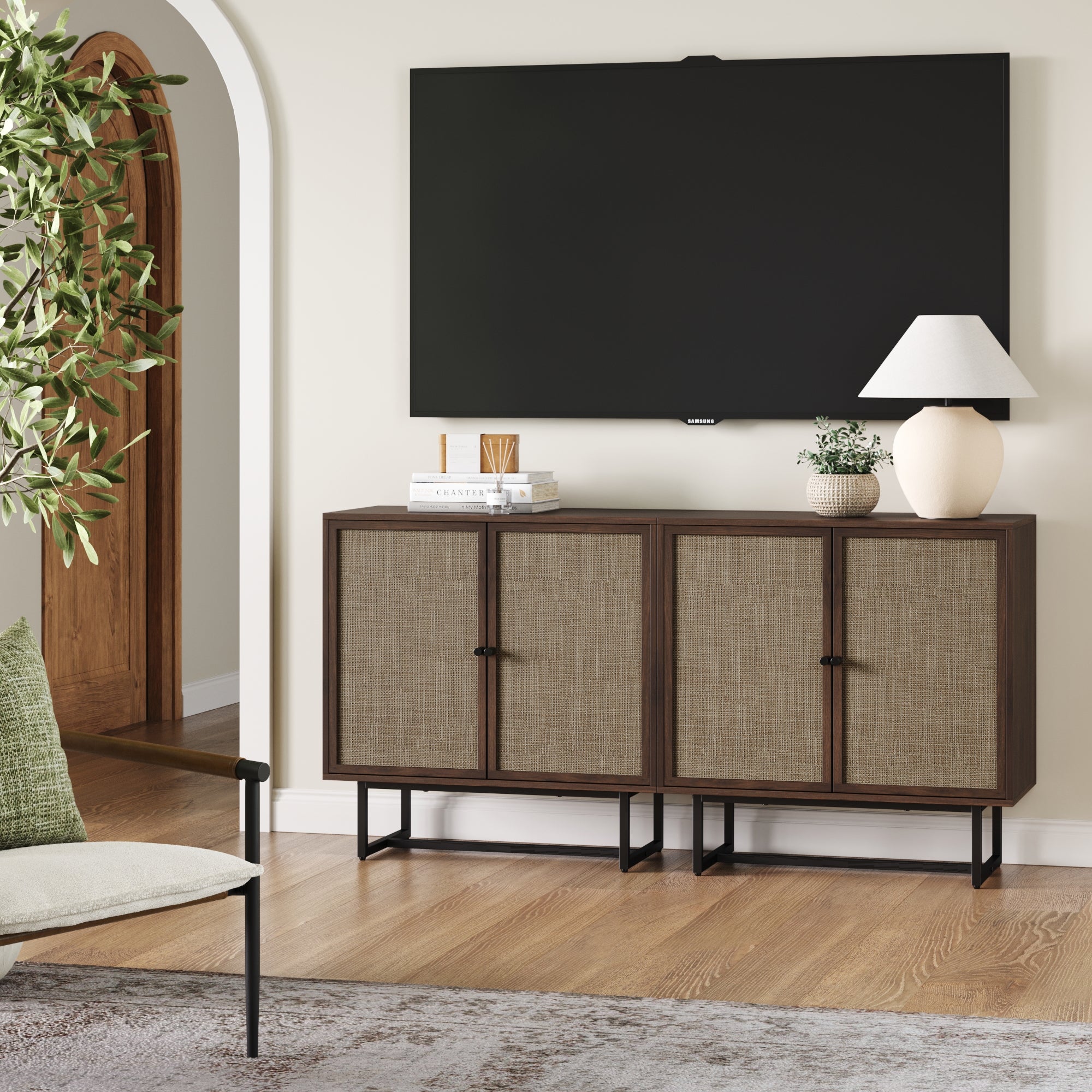 A modern living room features a large flat-screen TV mounted above two Nathan James Rattan TV Console Credenzas in walnut, displaying books, a lamp, and a vase. A green plant and cushioned chair are partially visible in the foreground.