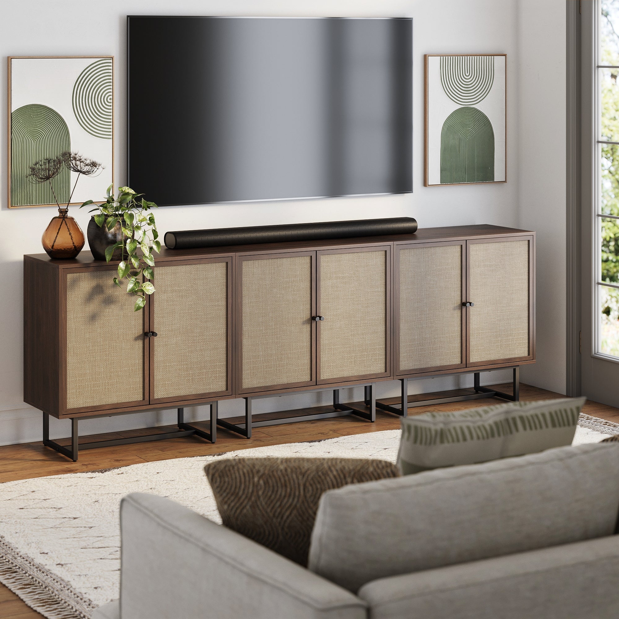 Modern living room with a large TV mounted above a long wooden sideboard, decorated with plants and minimalist art. A beige sofa and patterned rug are visible in the foreground. Sunlight streams through a window on the right.