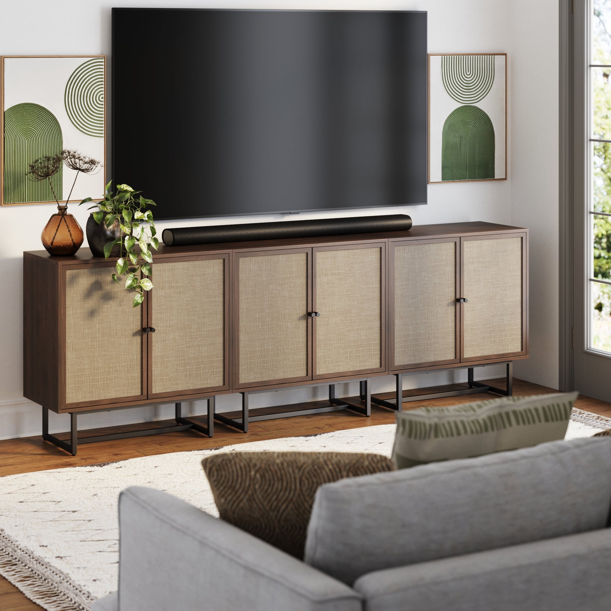 A modern living room showcases a large flat-screen TV on the Nathan James Rattan TV Console Credenza Walnut (Set of 3), with abstract green art, decorative vases, a plant, and a light sofa with textured pillows in the foreground.