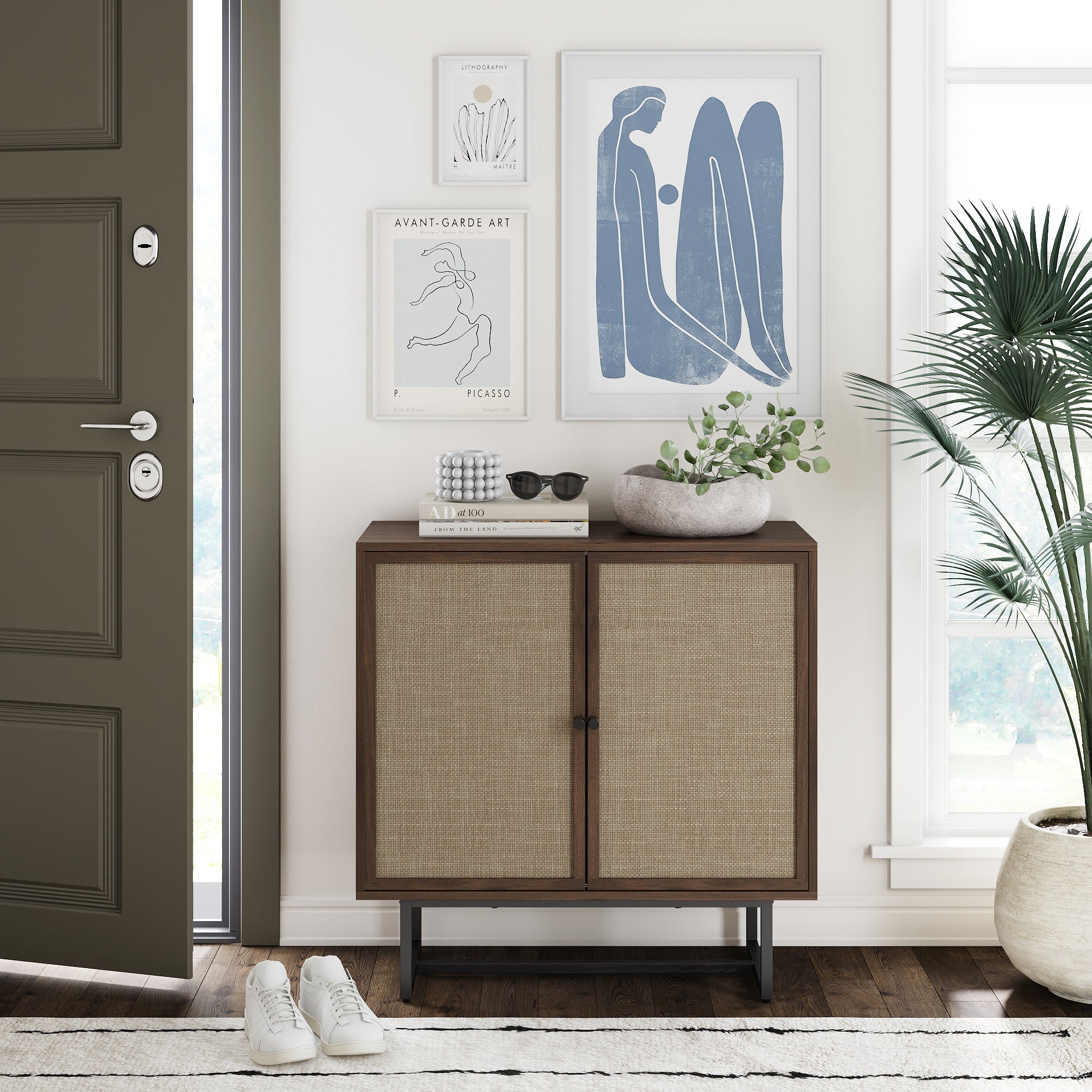 A modern entryway features the Nathan James Wood & Rattan Storage Cabinet in walnut with an adjustable shelf, books, sunglasses, a white bowl with greenery, framed abstract art, and white sneakers on the wooden floor by a beige door.