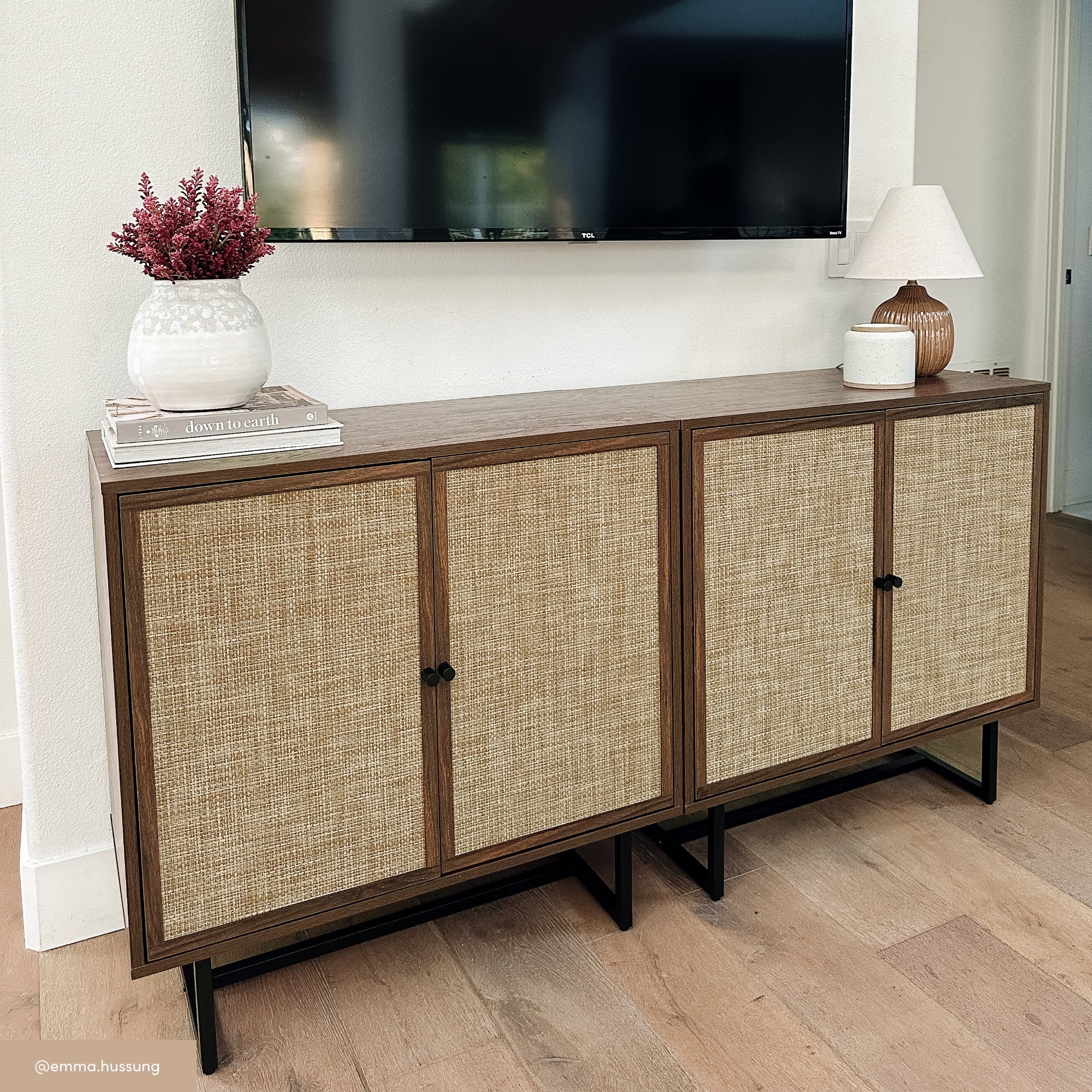 A wooden sideboard with woven rattan doors stands against a white wall beneath a mounted TV. On top are a lamp, a vase with pink flowers, and two stacked books. The floor is light wood.
