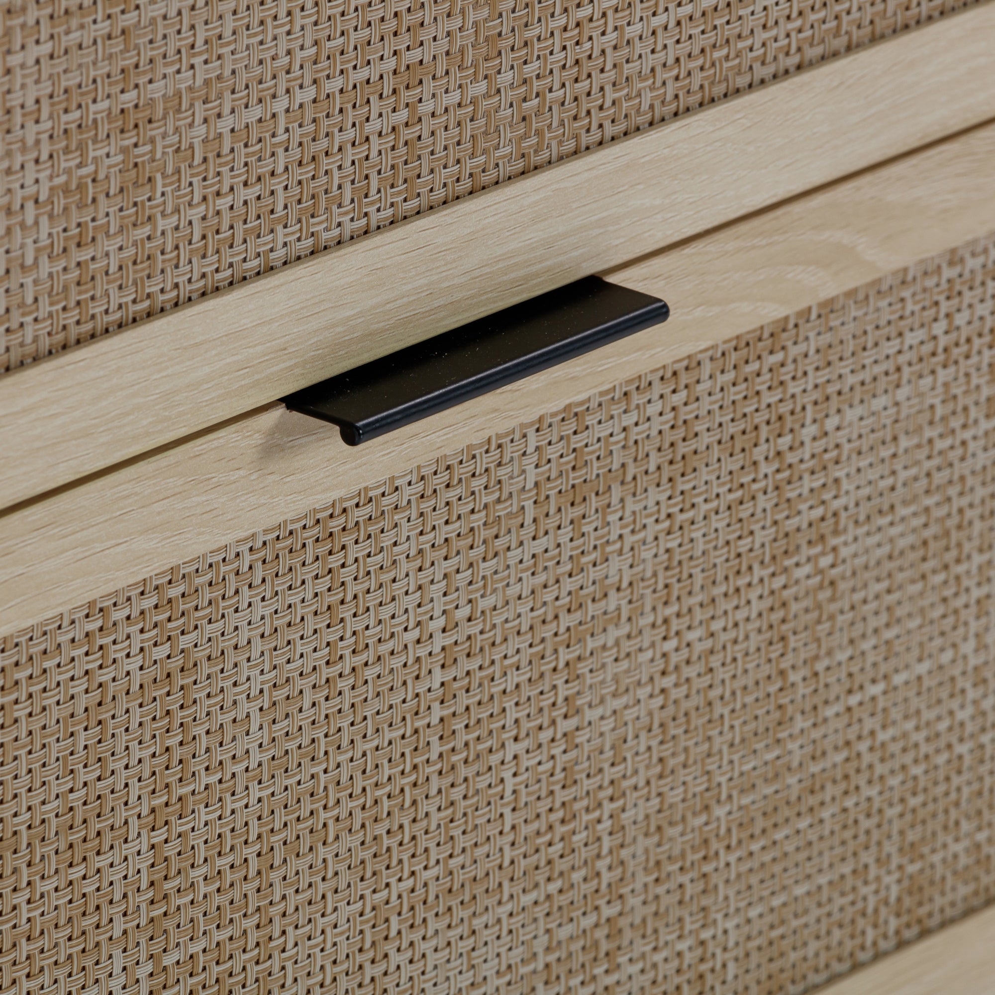 Close-up of a wooden drawer with a woven rattan front panel and a thin, black metal handle. The wood has a light, natural finish, and the woven texture adds a decorative element.