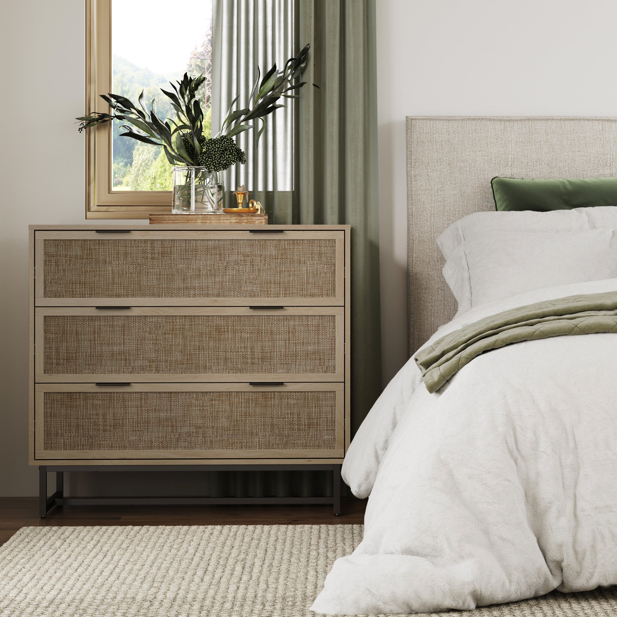A modern bedroom with a beige bed, green pillow, and blanket. Next to it is a wooden dresser with three drawers, topped with plants and decor. A window with green curtains lets in natural light.