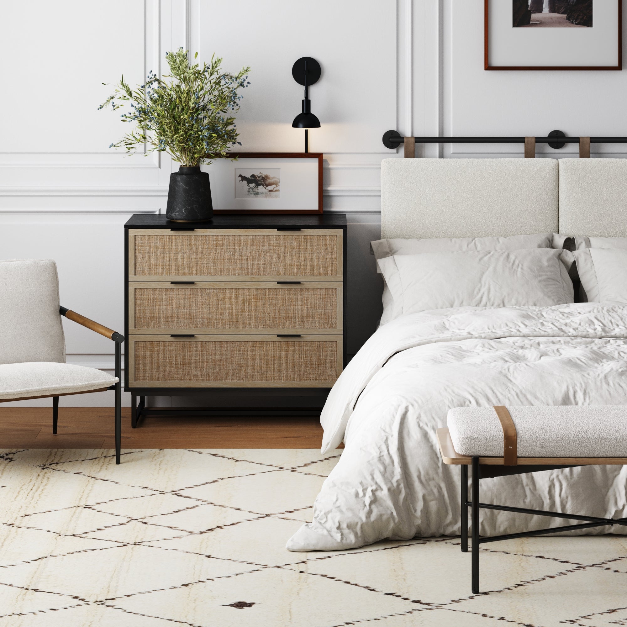 A modern bedroom featuring a light upholstered bed, white bedding, wooden nightstand, Nathan James Rattan, Wood & Metal 3-Drawer Dresser in black, potted plant, framed picture, wall sconce, cushioned bench, armchair, and patterned rug.