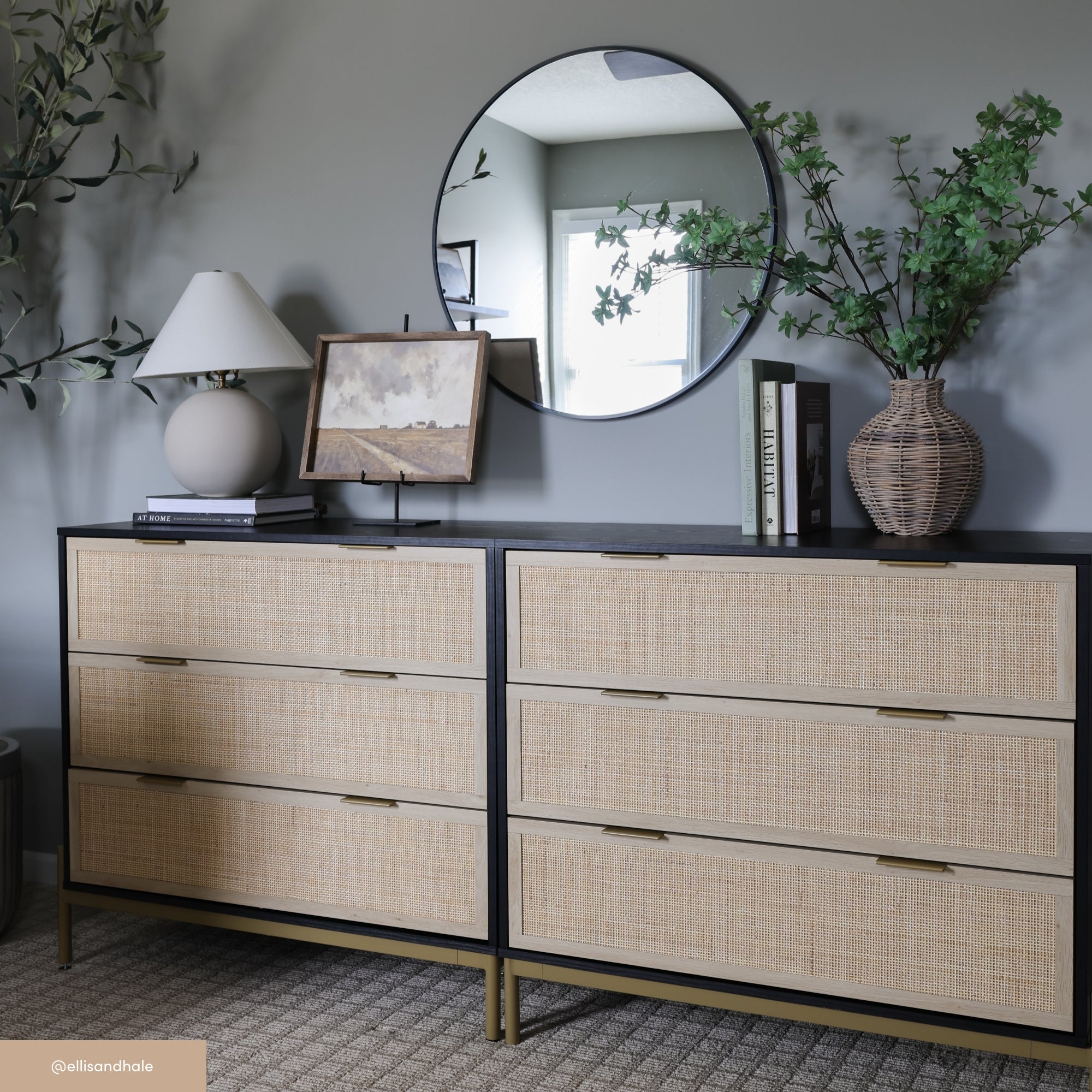 A modern dresser with woven drawer fronts, topped with a lamp, framed art, books, and a vase with greenery. A round mirror hangs above, reflecting part of the room. The walls are gray, and the floor is carpeted.
