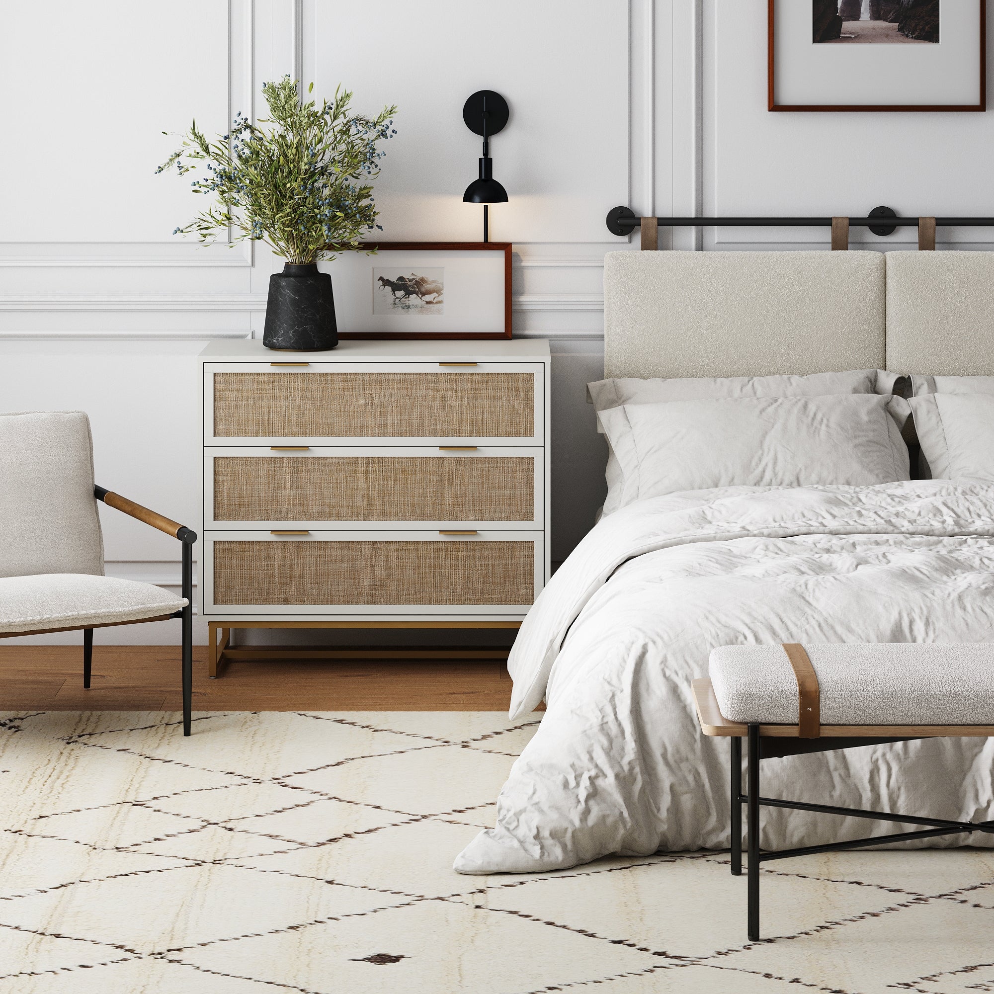 A modern bedroom with a beige bed, white bedding, cushioned bench, light armchair, and the Nathan James Rattan, Wood & Metal 3-Drawer Dresser in white. Decor includes a vase with greenery, wall art, and a black wall sconce.