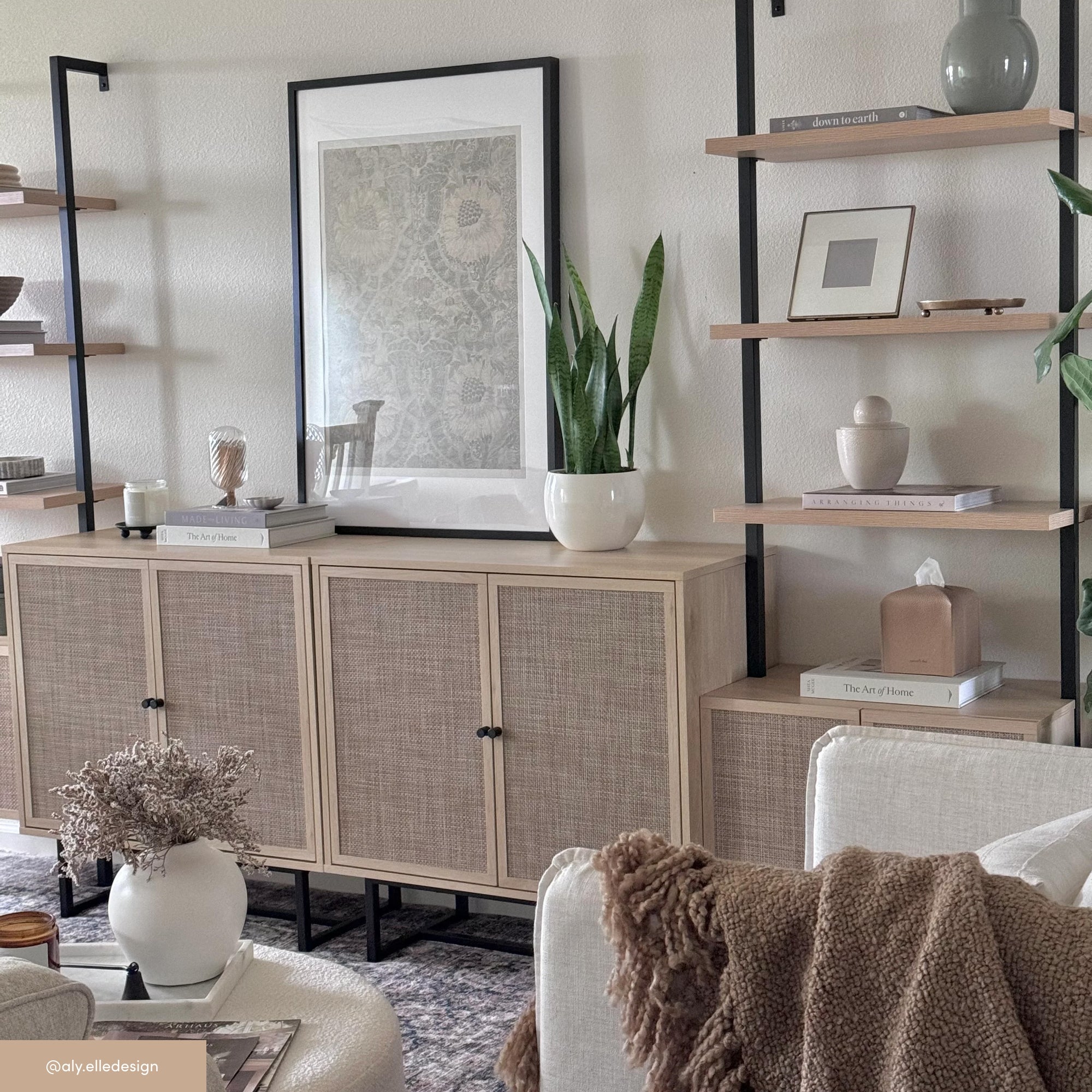 A modern living room features light wood cabinets, open shelves with decorative objects, a potted plant, framed artwork, stacked books, and neutral-toned furniture with cozy textures.