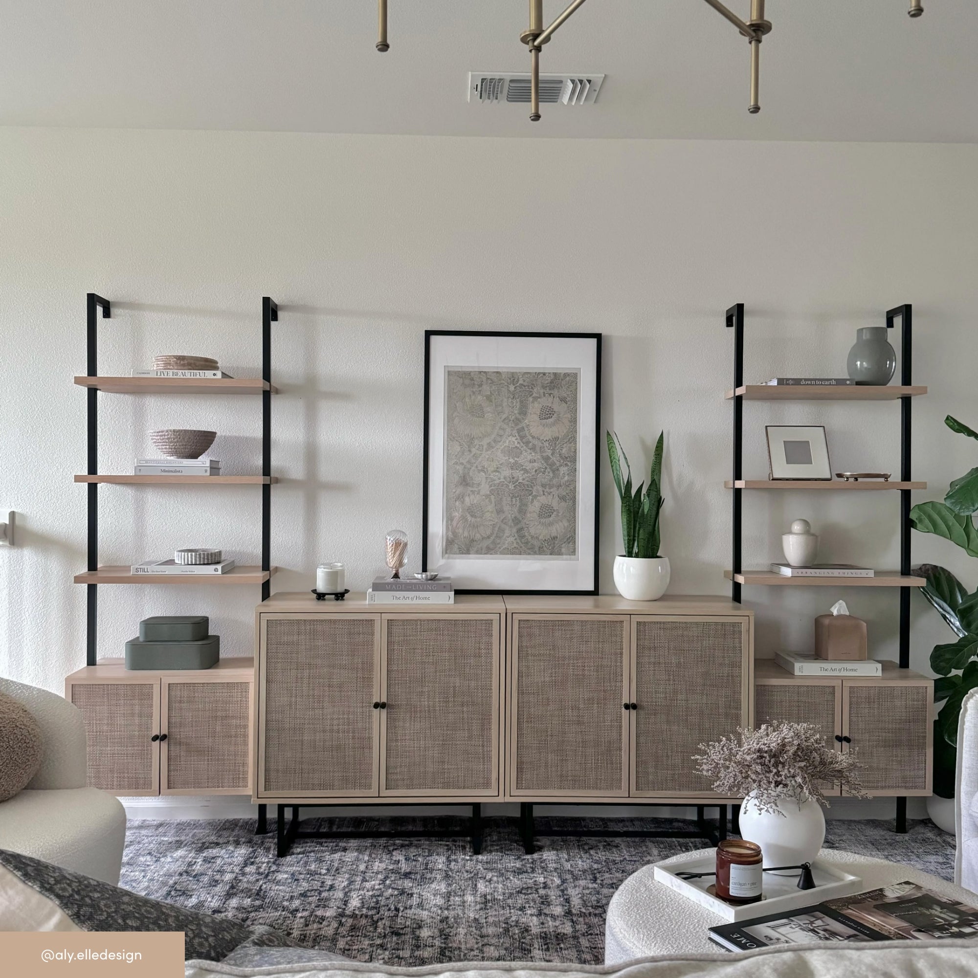A modern living room with a neutral color palette features two shelving units with decor items, a framed artwork, wicker cabinets, a patterned rug, and a round coffee table with a vase of flowers.