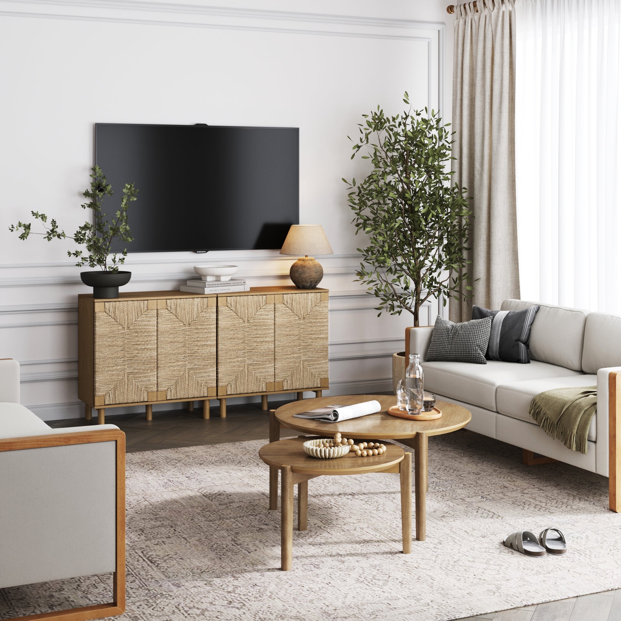 A modern living room with neutral tones, featuring a wall-mounted TV, a wooden cabinet, two round nested coffee tables, light sofas, potted plants, and soft natural light coming through beige curtains.