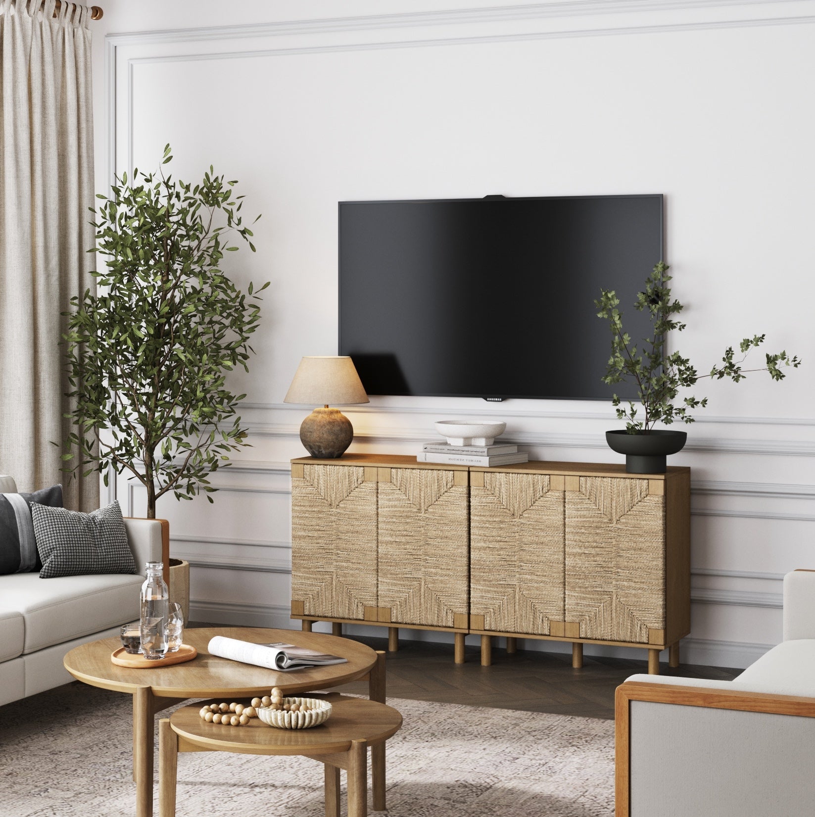 A modern living room with a beige sofa, wooden coffee table, textured sideboard with lamp and plant, large flat-screen TV on the wall, and tall plants in the corners. Neutral tones and natural light create a cozy atmosphere.