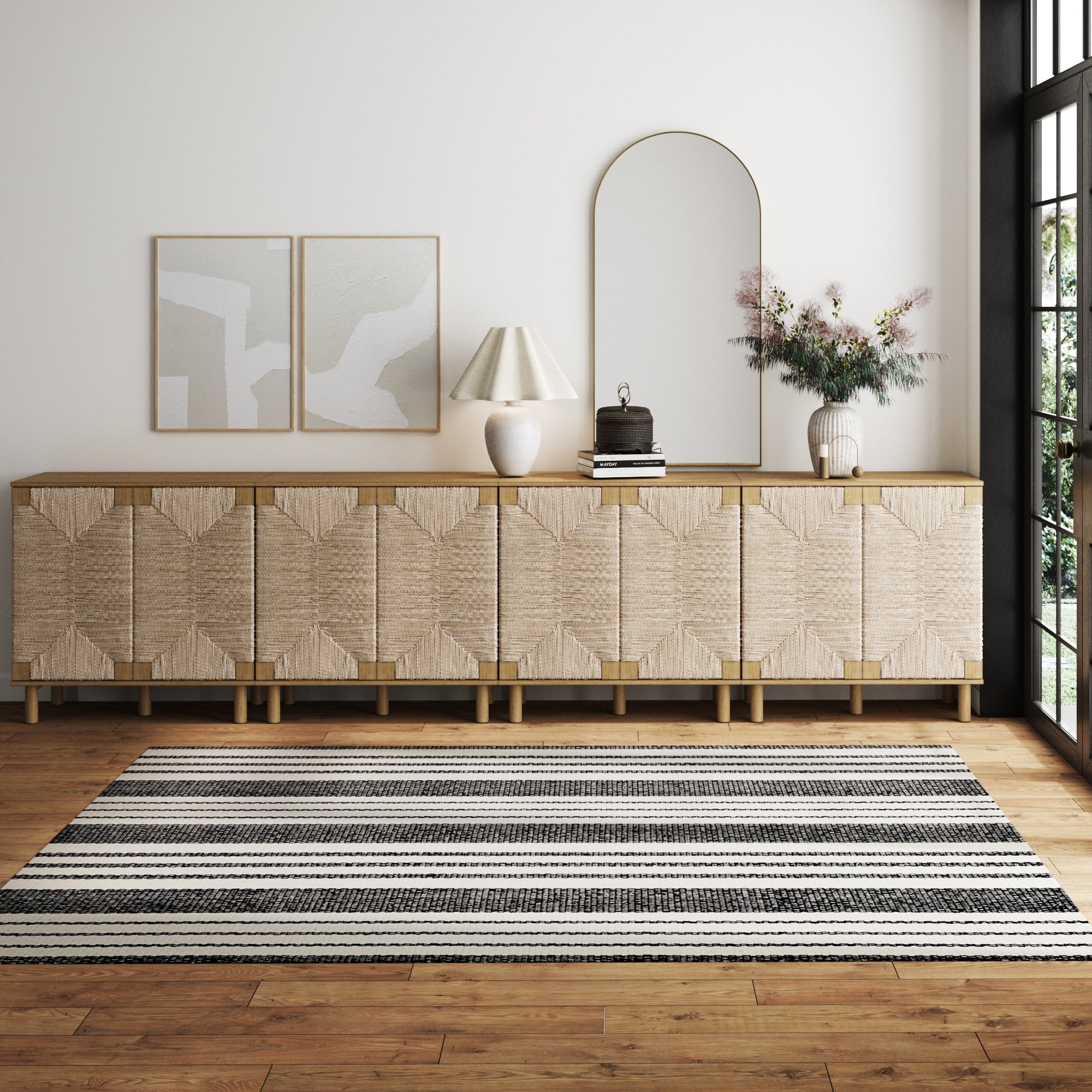 A modern living room features a woven sideboard, two abstract art pieces, a large arched mirror, a lamp, books, a vase with flowers, and a black-and-white striped rug on a wooden floor.
