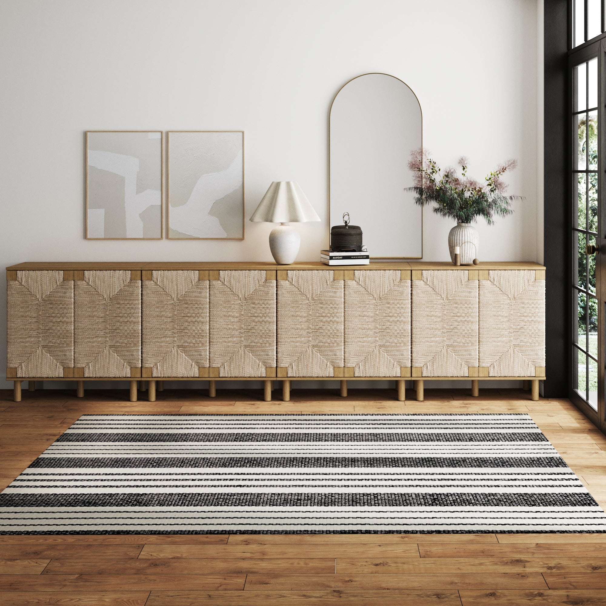 A modern, minimal living room features the Nathan James Large Seagrass TV Console Credenza (set of 4), two abstract framed artworks, a round mirror, lamp, books, vase with flowers, and a black-and-white striped rug on a wooden floor.