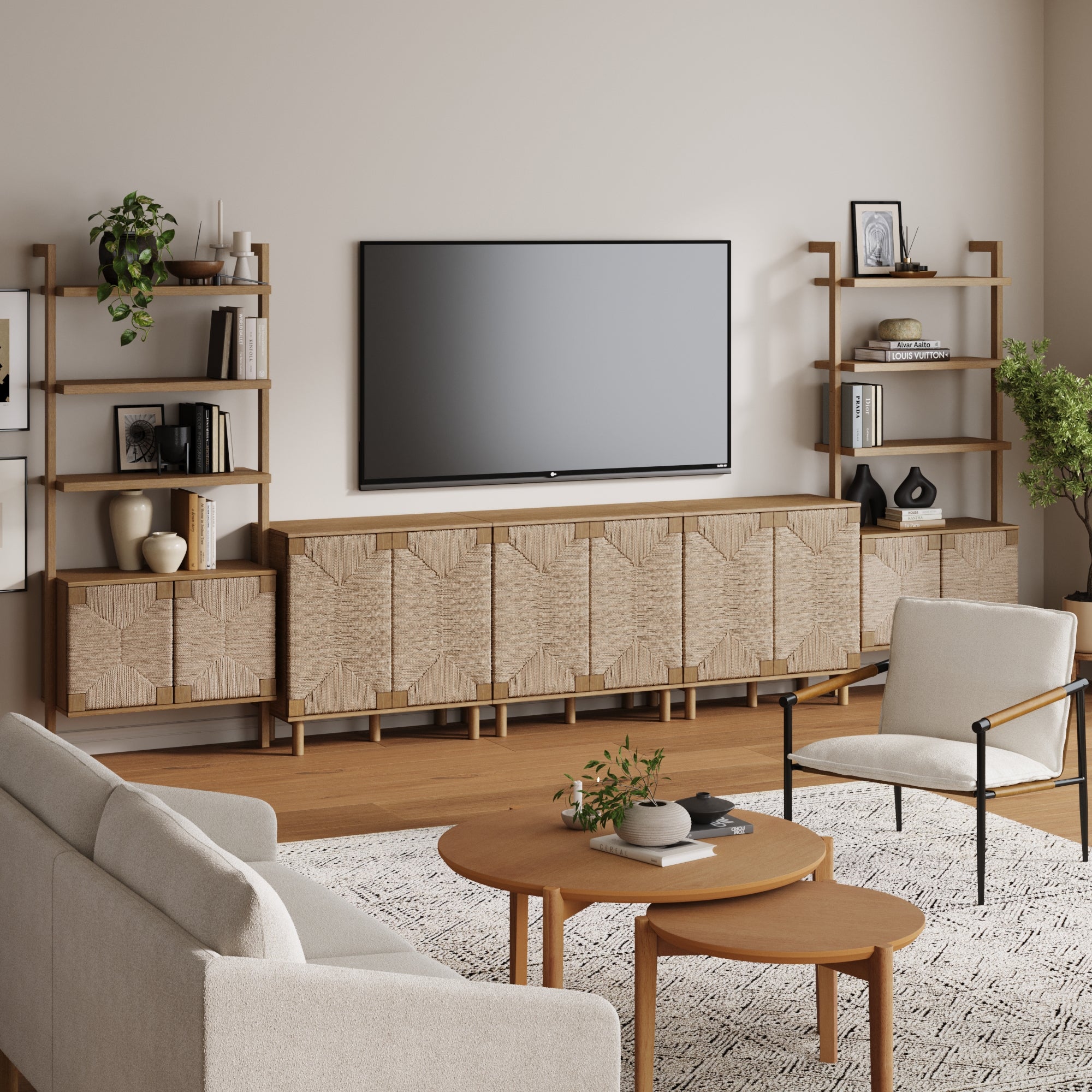 A modern living room featuring the Nathan James Boho Seagrass Entertainment Center in light brown with three cabinets, neutral armchairs, a beige sofa, round wooden coffee tables, a patterned rug on wood floors, plus plants, books, and vases.