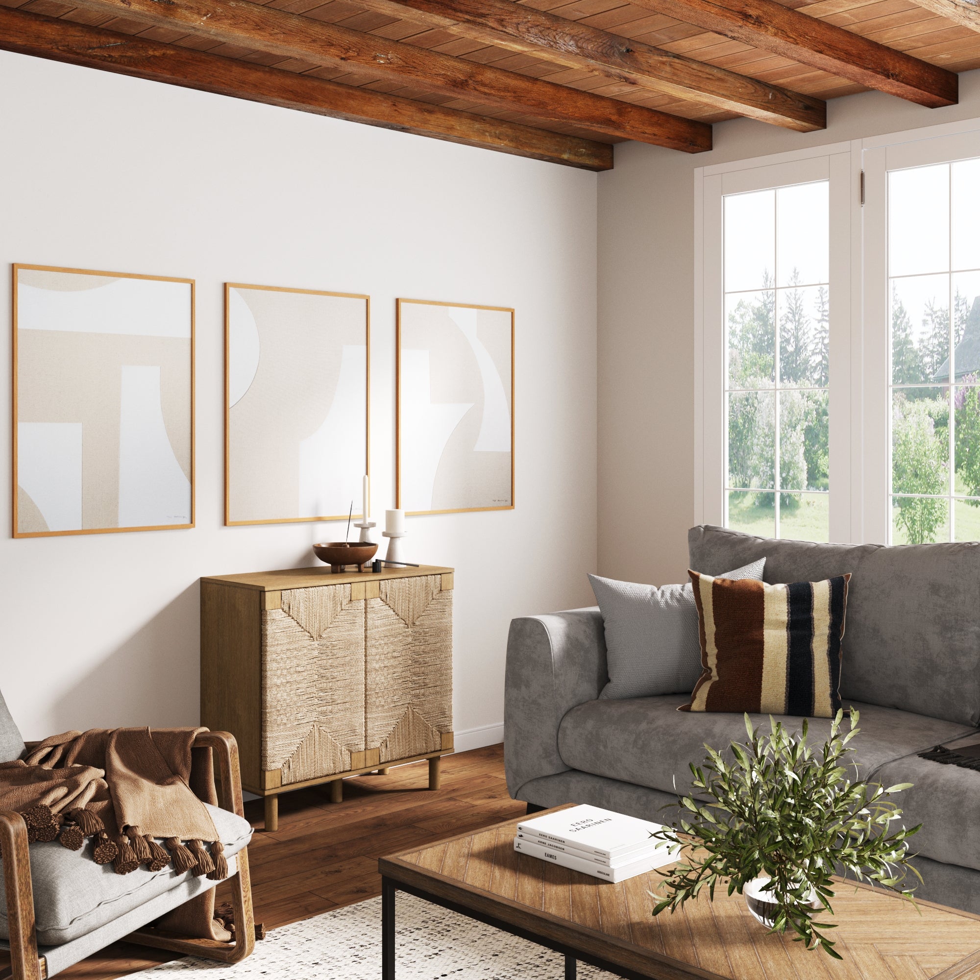 A cozy living room with a gray sofa, wooden coffee table, striped pillow, and brown throw blanket. Three abstract art pieces hang above a wooden cabinet. Large windows let in natural light and show a green outdoor view.