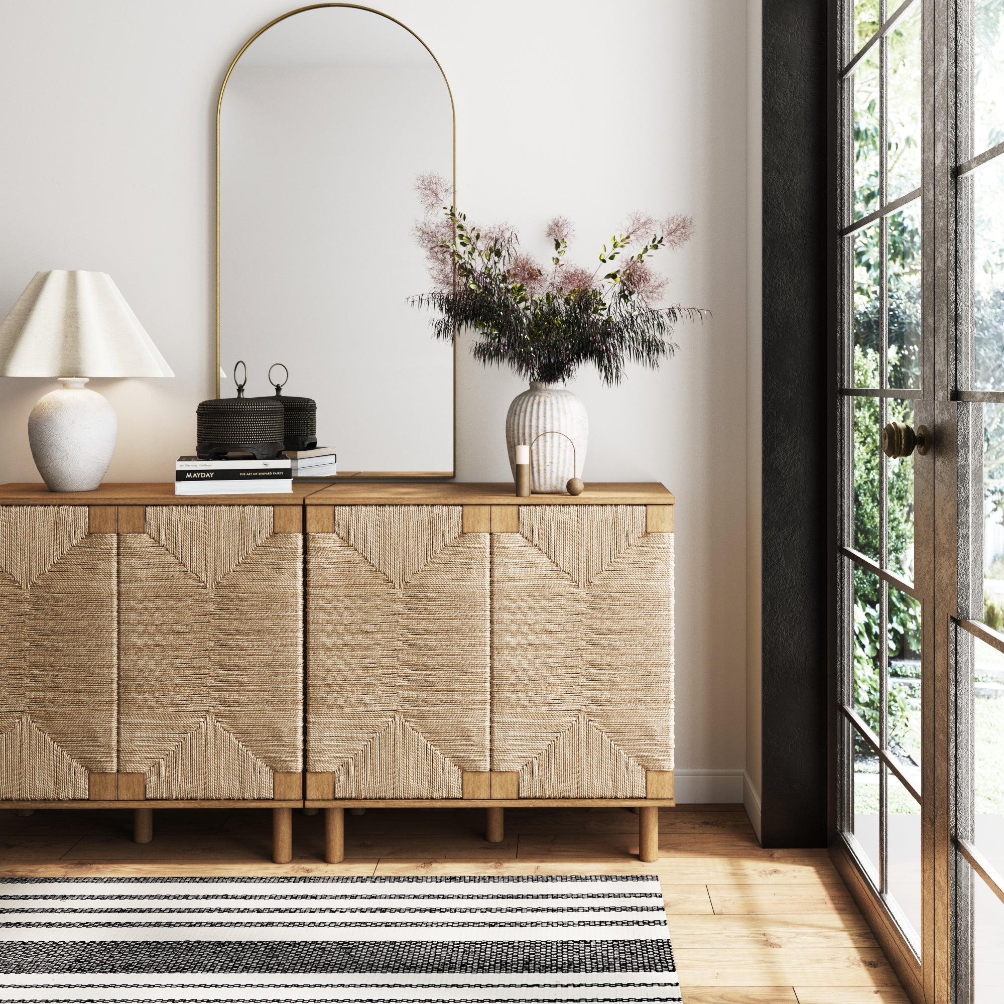 A modern console table with a textured front, topped with a white lamp, books, black decor, and a vase of flowers. A tall, arched mirror hangs above. Sunlight streams in from large glass doors beside a striped rug.