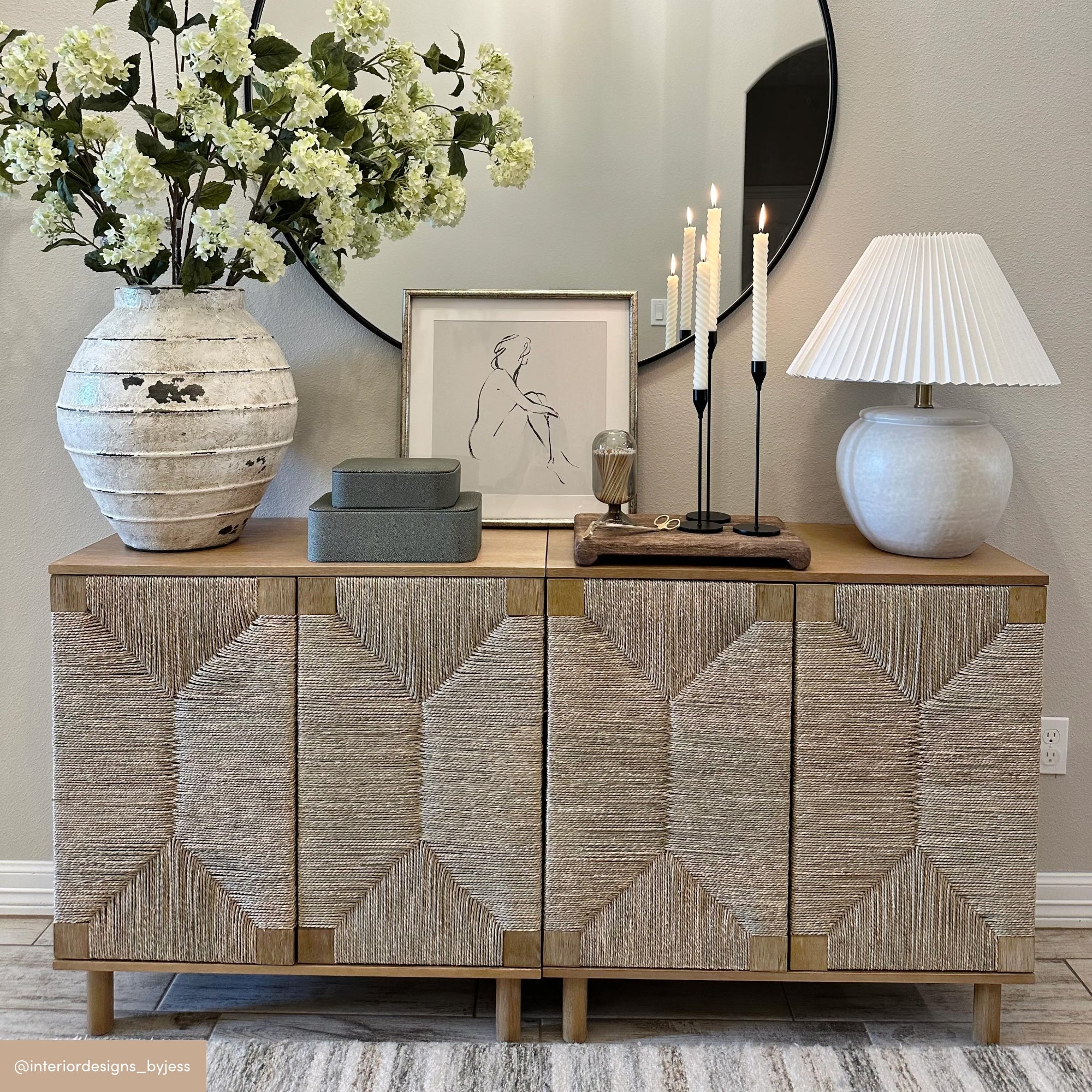 A modern sideboard with textured doors holds a large ceramic vase of white flowers, a framed line art portrait, stacked boxes, tall candles, and a white pleated lamp. A round mirror hangs on the beige wall above.