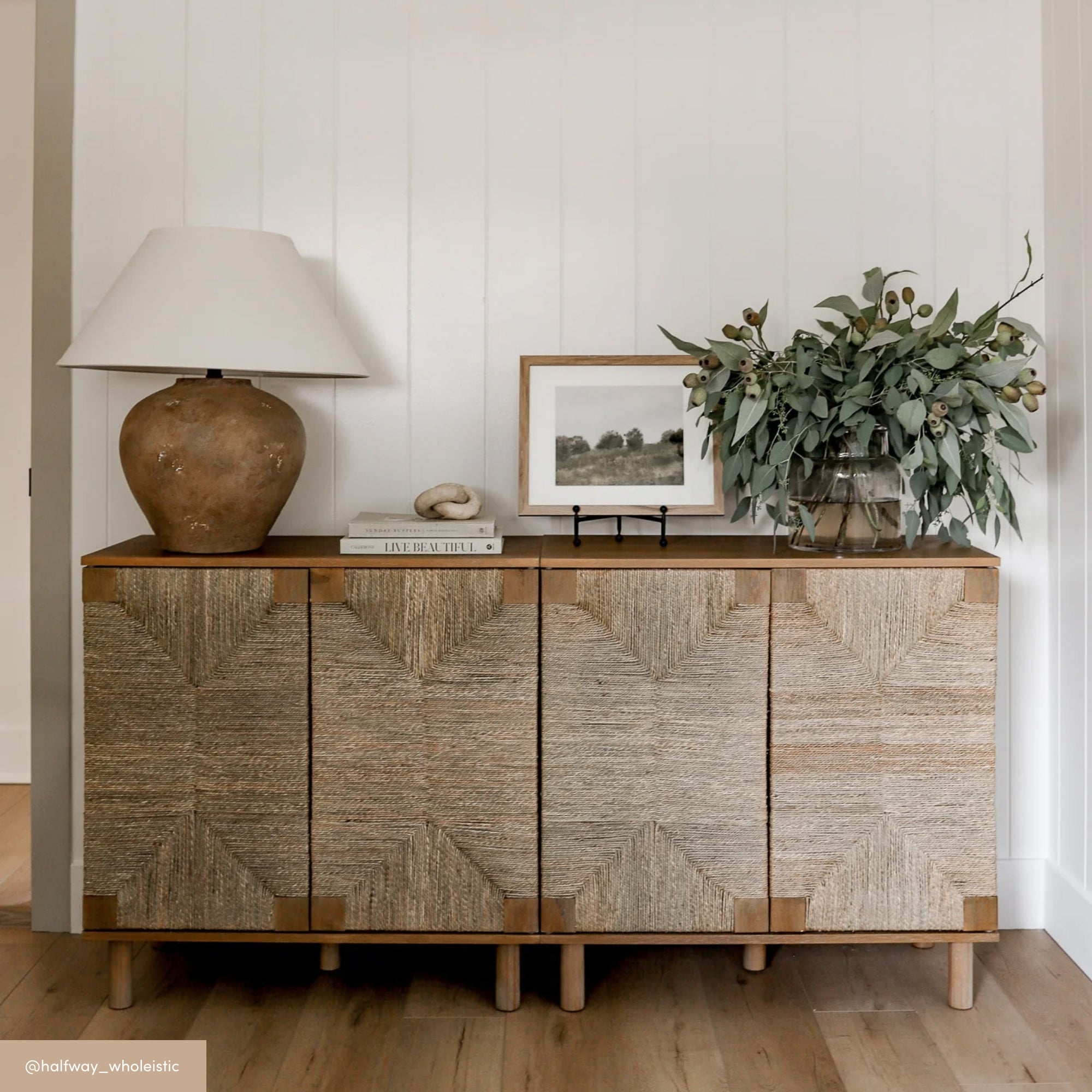 A textured sideboard with a brown lamp, books, framed art, and a glass vase of greenery sits against a white paneled wall on a light wood floor in a modern, minimalist room.
