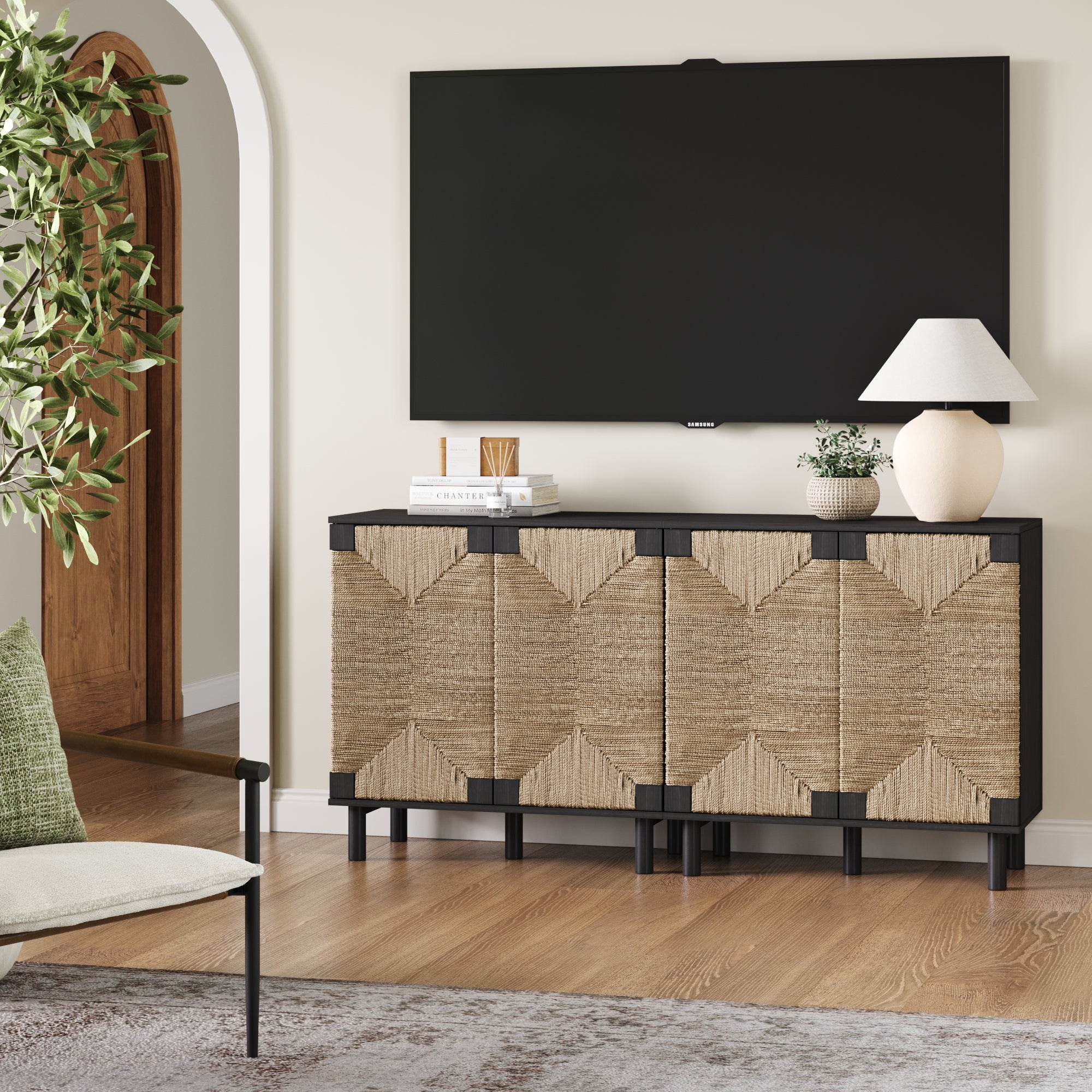 A modern living room showcases a flat-screen TV above the Nathan James Seagrass TV Console Credenza in Black (Set of 2), featuring natural seagrass cabinet doors. Books, a plant, vase, and lamp complete the chic bohemian decor.
