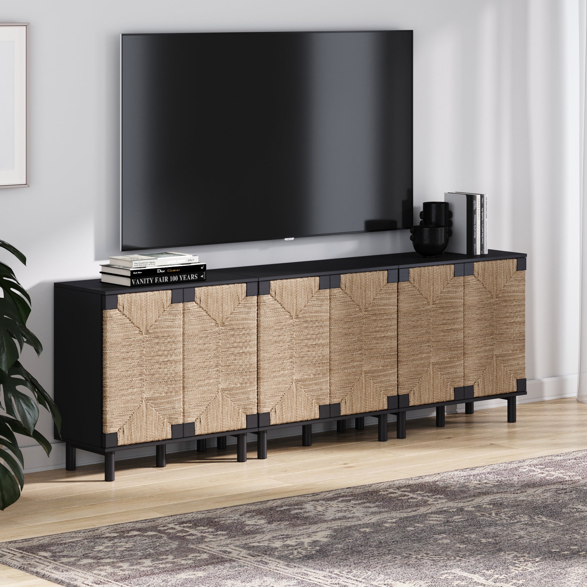 A modern living room showcases the Nathan James Seagrass TV Console Credenza Black (Set of 3), featuring woven rattan doors, topped with books and a vase, all set by a patterned rug on light wood floors for a bohemian touch.