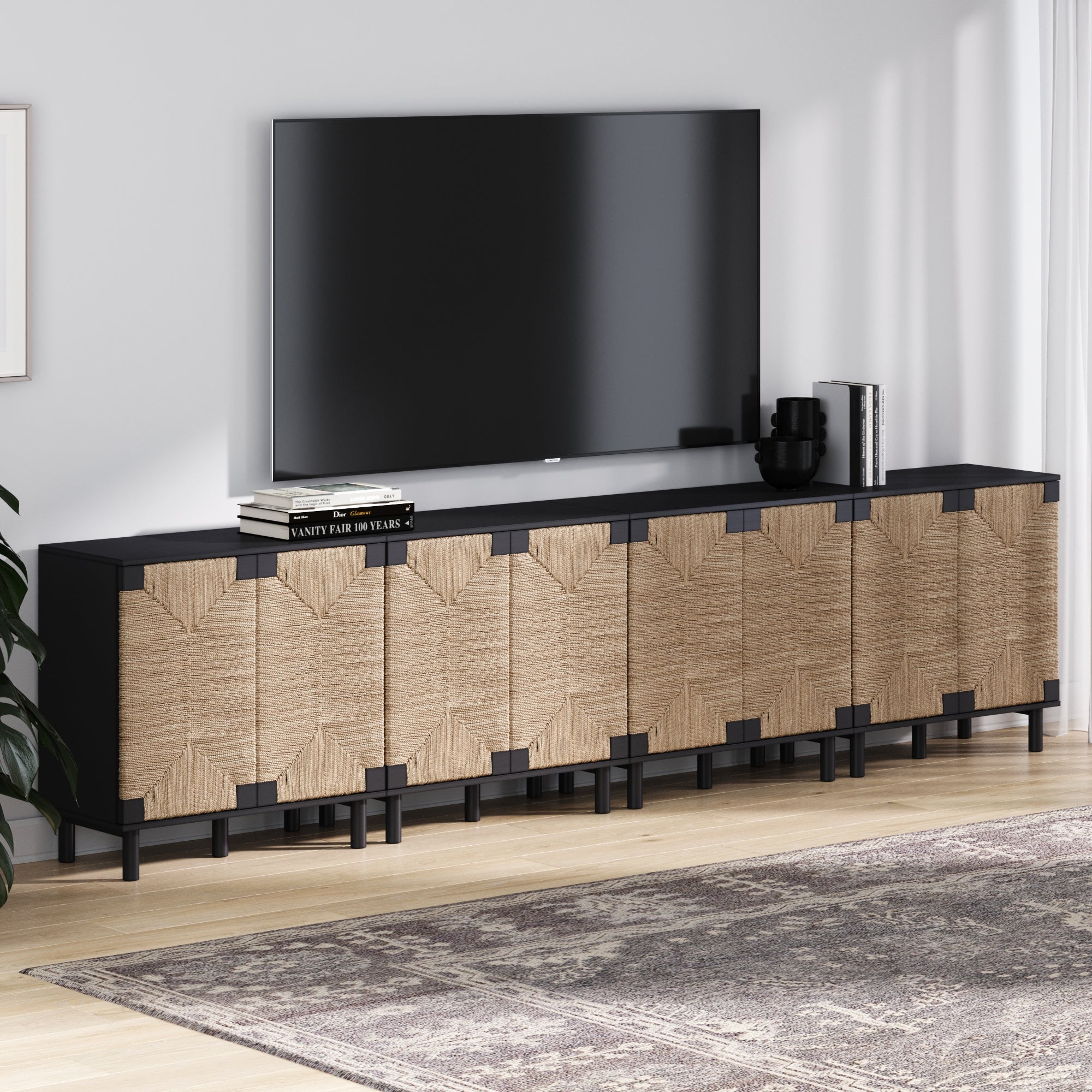 The Nathan James Large Seagrass TV Console Credenza Black (Set of 4) stands against the wall under a large TV, topped with decorative items and books, while a patterned rug lies over the light wood floor.