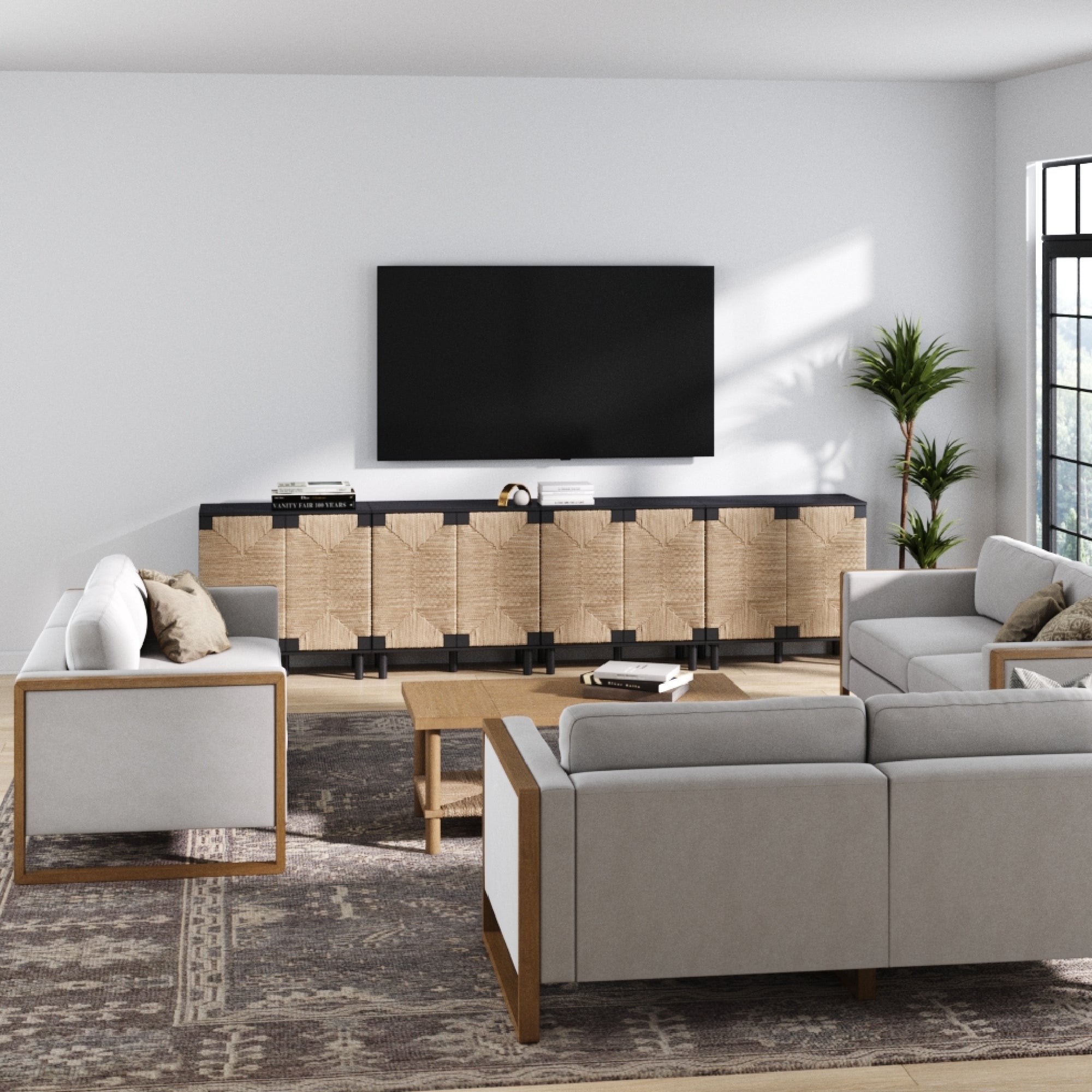 A modern living room with light gray sofas, a patterned rug, a wooden coffee table, and a large TV on the Nathan James Large Seagrass TV Console Credenza Black (Set of 4). A potted plant sits by the sunny window.