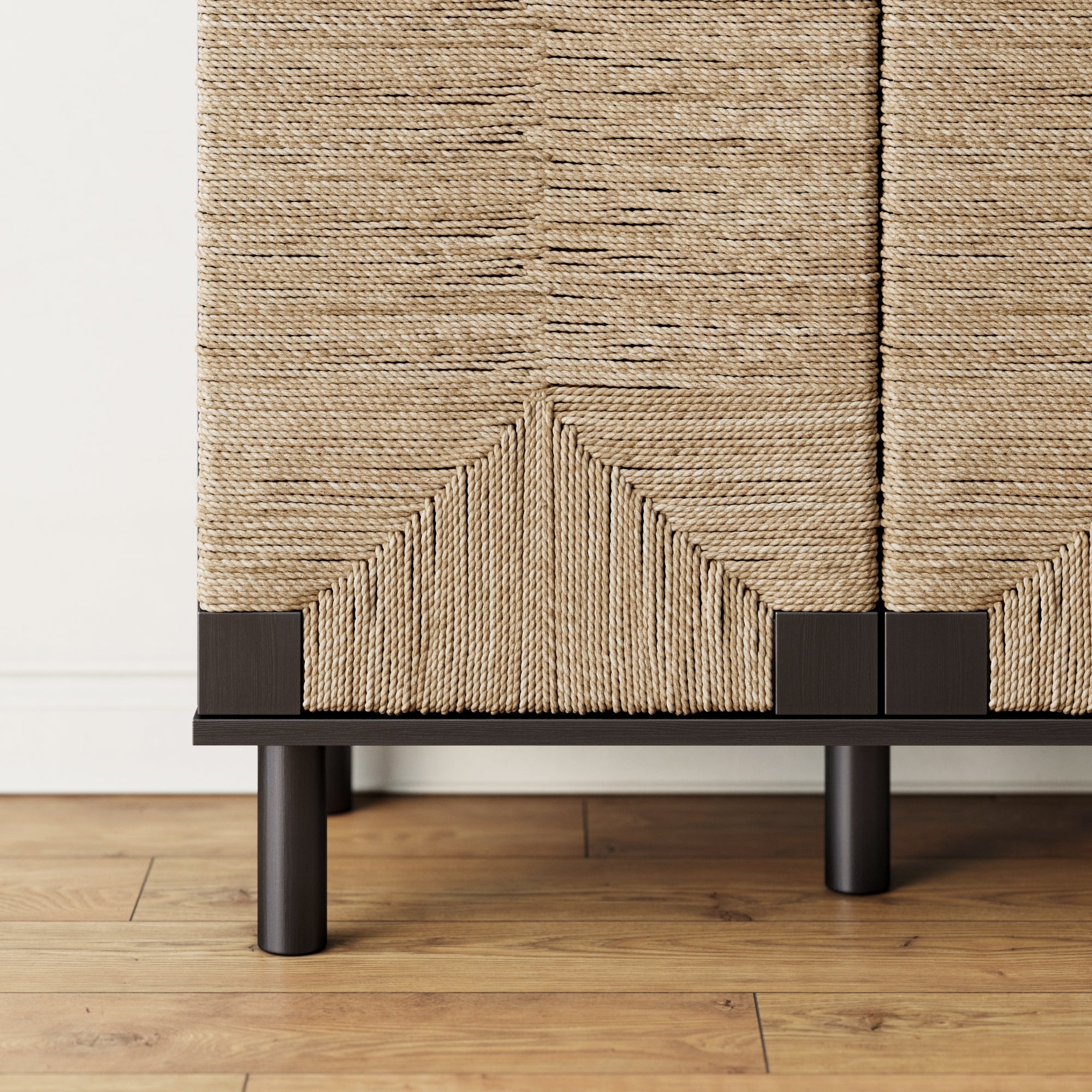 A close-up of a cabinet with textured, woven rattan doors and dark wood or metal legs, standing on a light wood floor against a white wall. The woven pattern forms geometric shapes at the base.