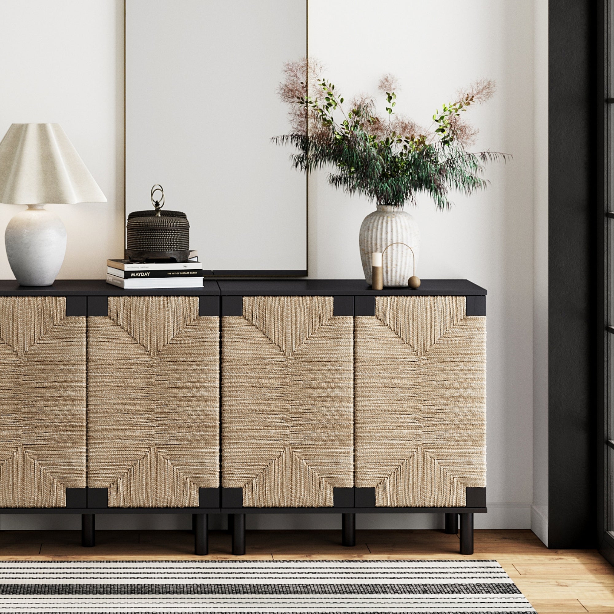 The Nathan James Seagrass TV Console Credenza Black (Set of 2) features natural seagrass cabinet doors and stands against a white wall, topped with a lamp, books, decor accents, and a vase; a mirror above adds bohemian style flair.