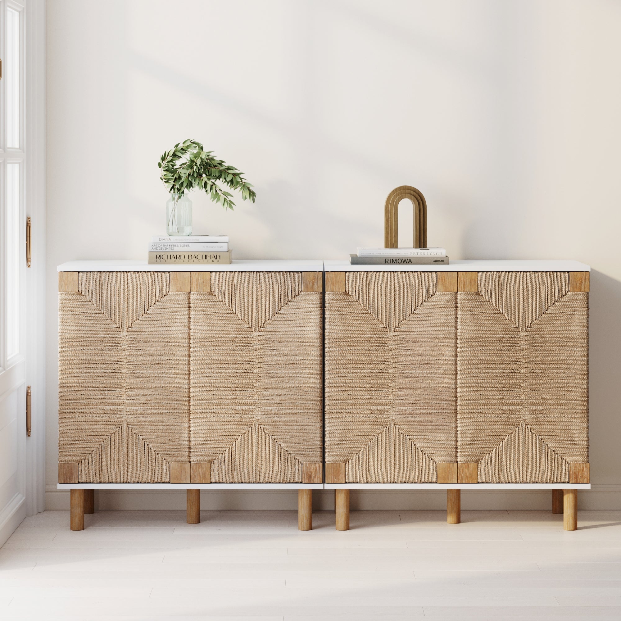 The Nathan James Seagrass TV Console Credenza White (Set of 2) features woven, textured doors and wooden legs, styled with a glass vase of greenery and a gold arch on top as sunlight pours in from the left window.