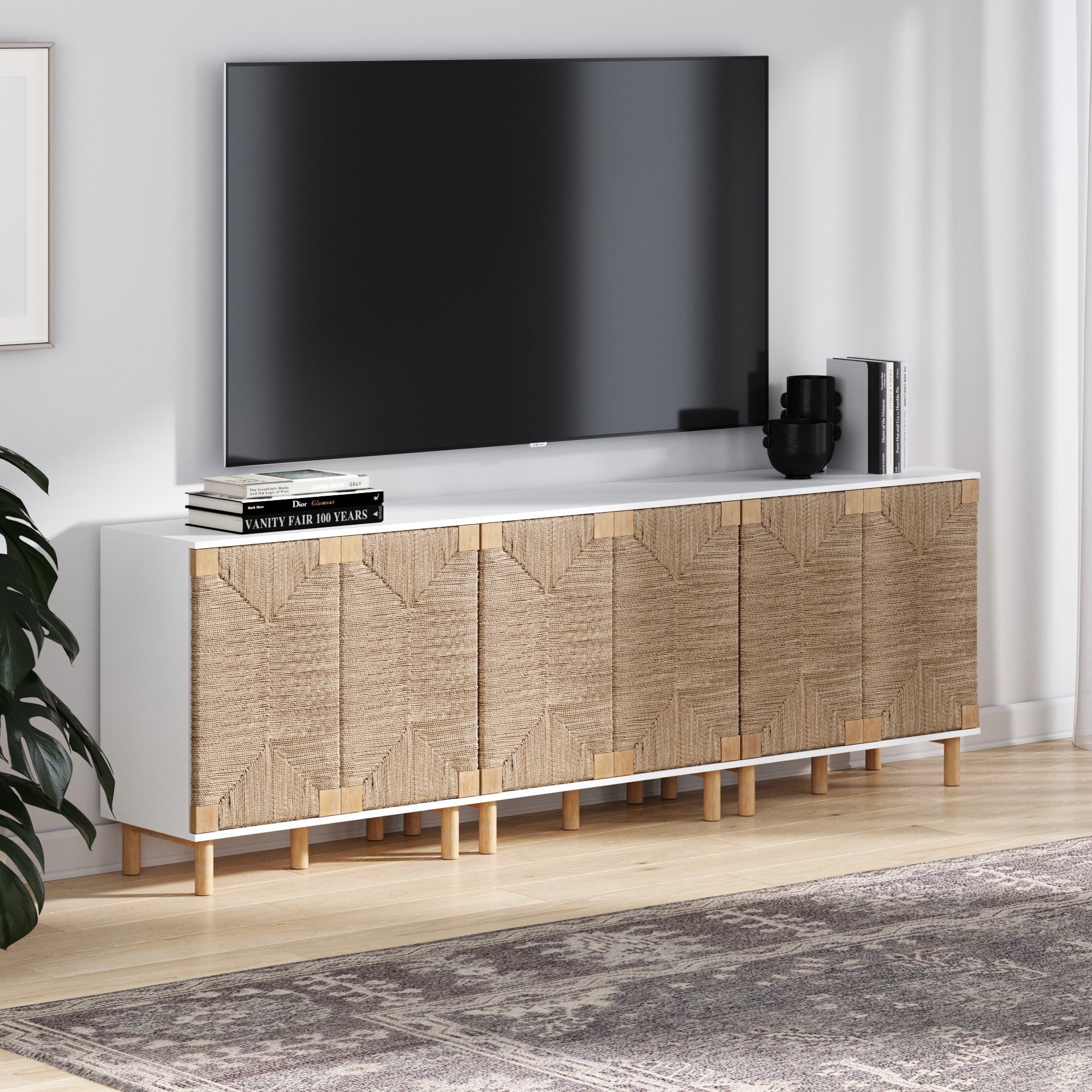 A modern living room with a large TV on the Nathan James Seagrass TV Console Credenza White (Set of 3), featuring woven doors, decorative books, a black vase, and a neutral patterned rug on wood floors.