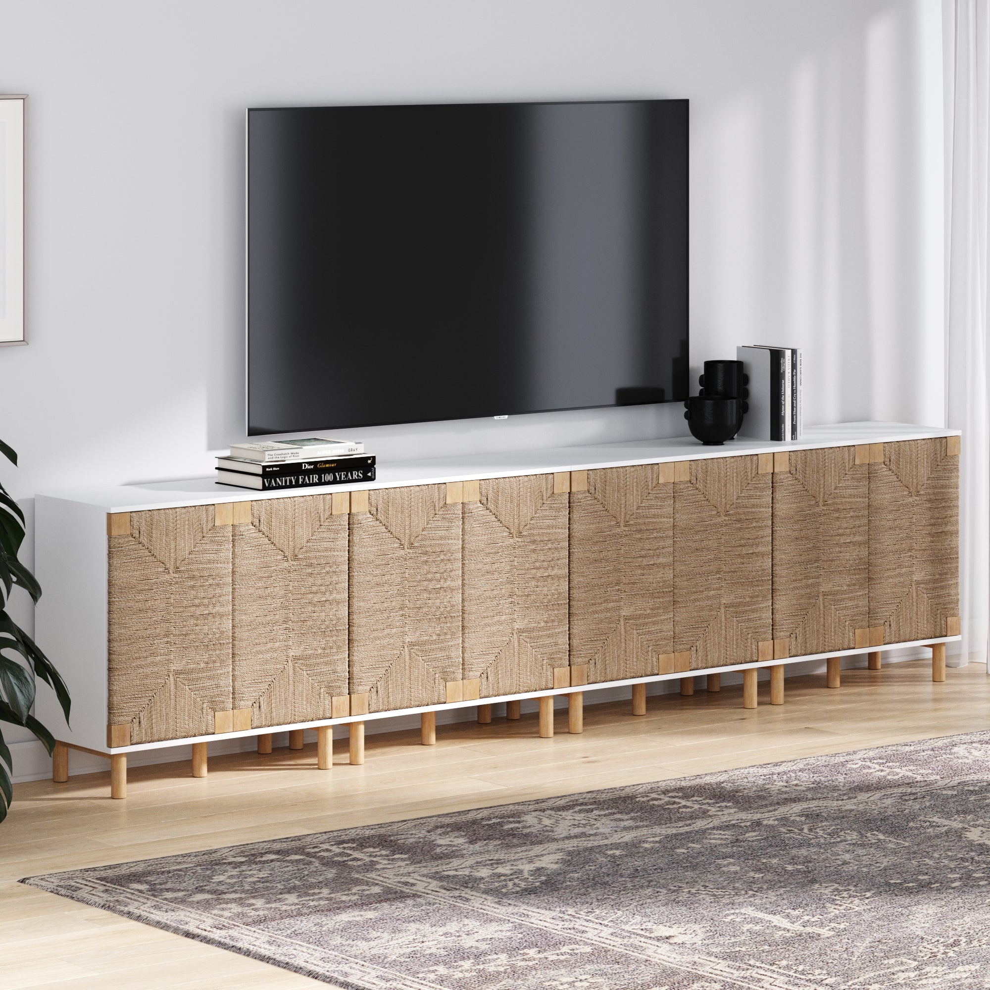 The Nathan James Large Seagrass TV Console Credenza White (Set of 4) features textured cabinet doors, a white top, and light wood legs. It holds a TV, books, and black decor on a light wood floor next to a patterned area rug.