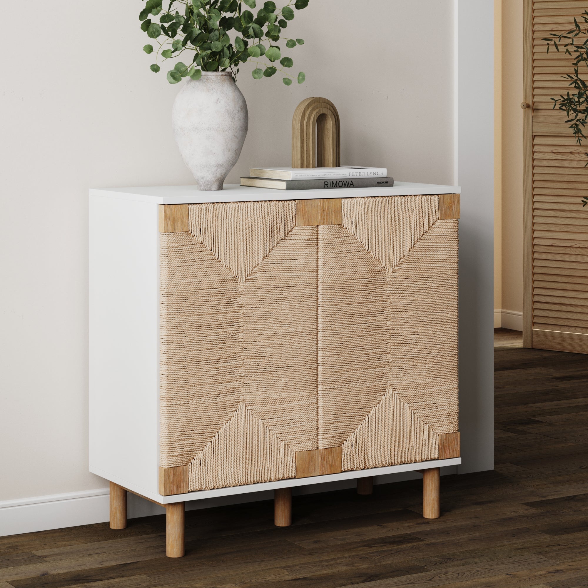 The Nathan James Wood & Seagrass Storage Cabinet in black stands on wooden legs, topped with a white ceramic vase, greenery, and décor for a boho look against light walls and wood flooring.