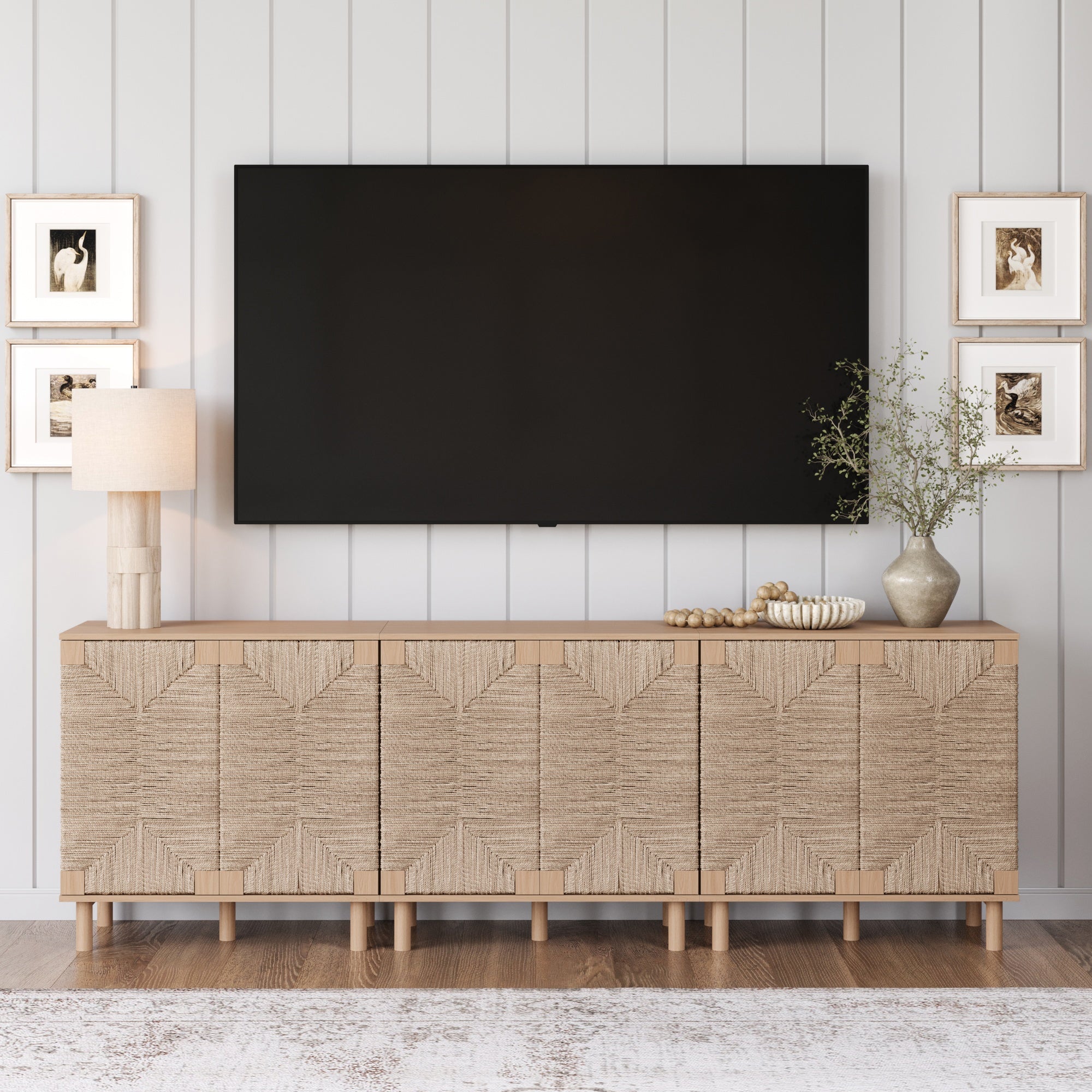 A modern living room showcases the Nathan James Seagrass TV Console Credenza Raw Oak (Set of 3) below a large wall-mounted TV, topped with a lamp, a vase of greenery, and a bowl, all framed by four pieces of art.