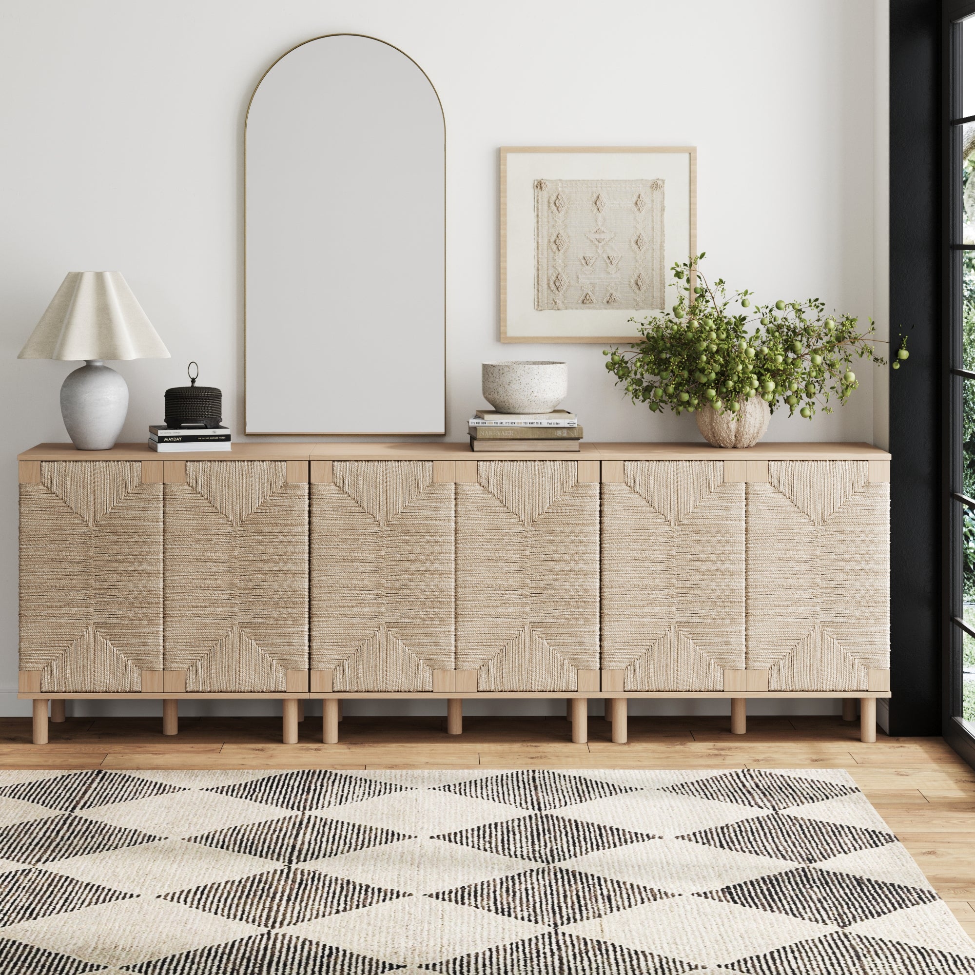 A modern living room showcases the Nathan James Seagrass TV Console Credenza Raw Oak (Set of 3) with natural seagrass doors, paired with a large arched mirror, table lamp, books, bowls, leafy plant, wall art, and a geometric rug.