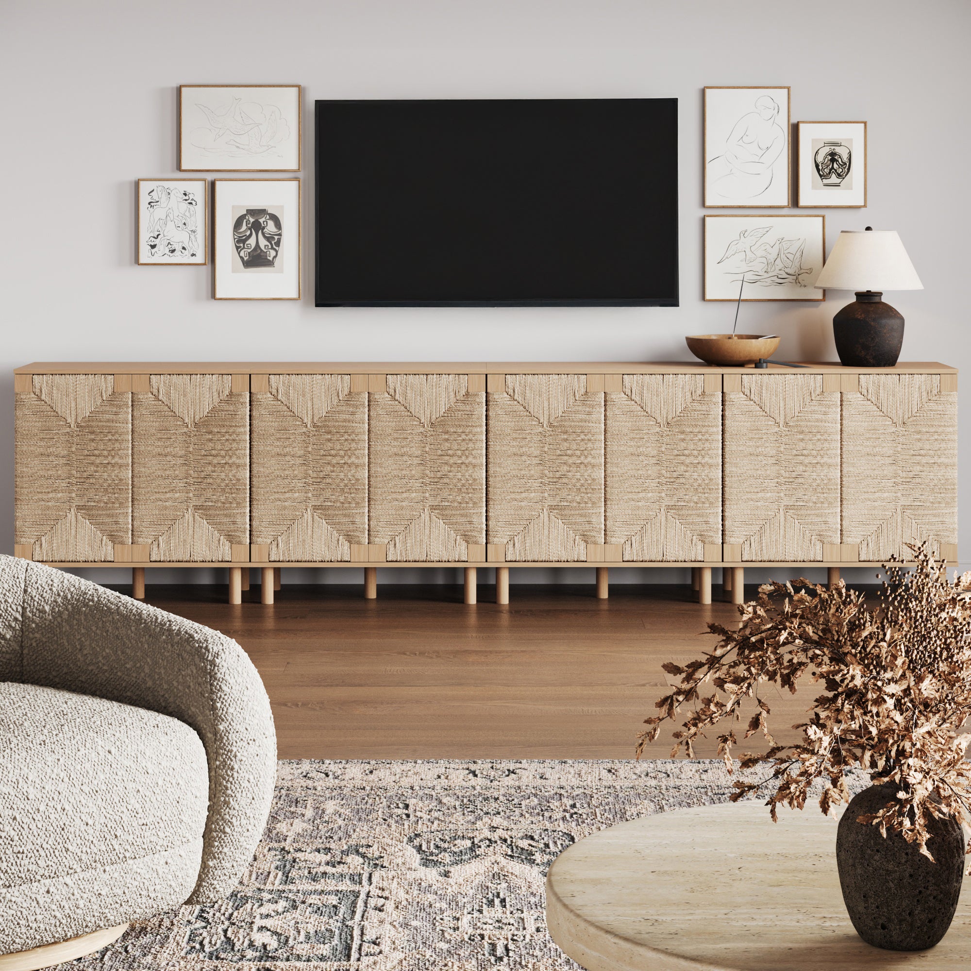 A modern living room with the Nathan James Large Seagrass TV Console Credenza in raw oak (set of 4), natural seagrass doors, a large black TV above, framed art, a lamp and bowls on top, round coffee table with dried flowers, and a curved armchair.