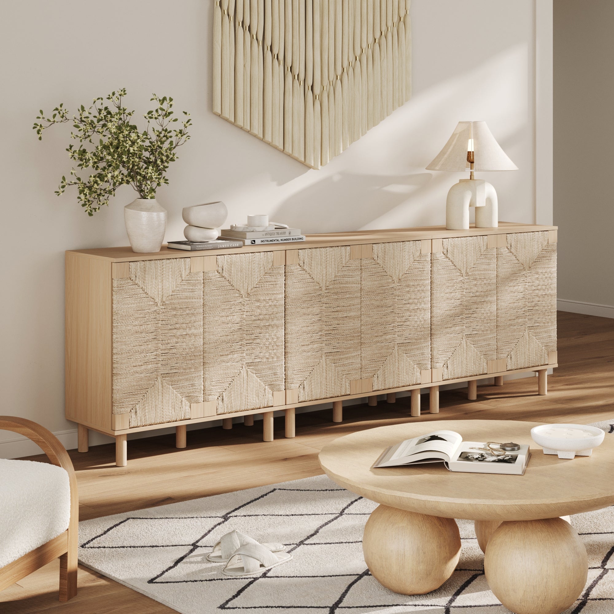 A modern living room showcases the Nathan James Wood & Seagrass Storage Cabinet in Raw Oak with light wood doors, decorative ceramics, a lamp, and a plant. A round coffee table, open book, and soft rug enhance the minimalist, boho-inspired decor.