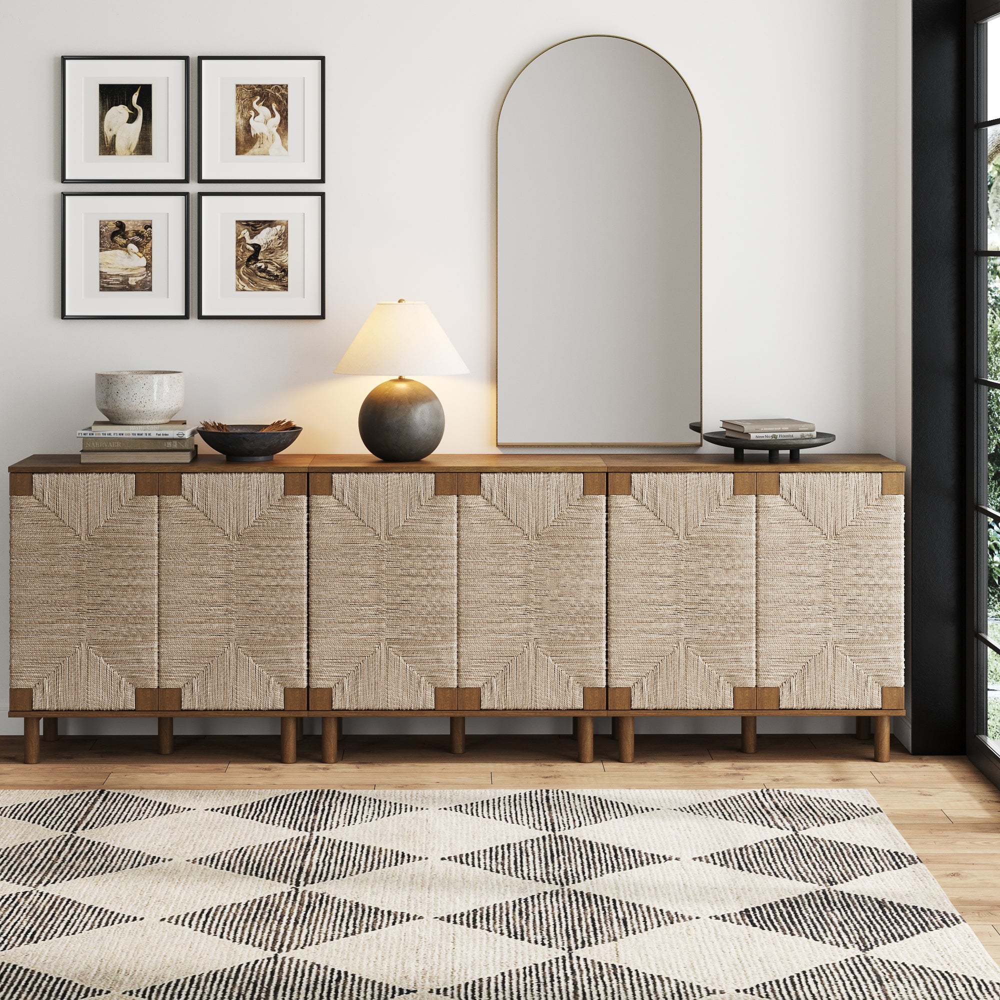 A modern living room with the Nathan James Seagrass TV Console Credenza in dark brown (set of 3), natural seagrass doors, a table lamp, decorative bowls, books, an arched mirror, four framed prints on the wall, and a geometric rug on wood floors.