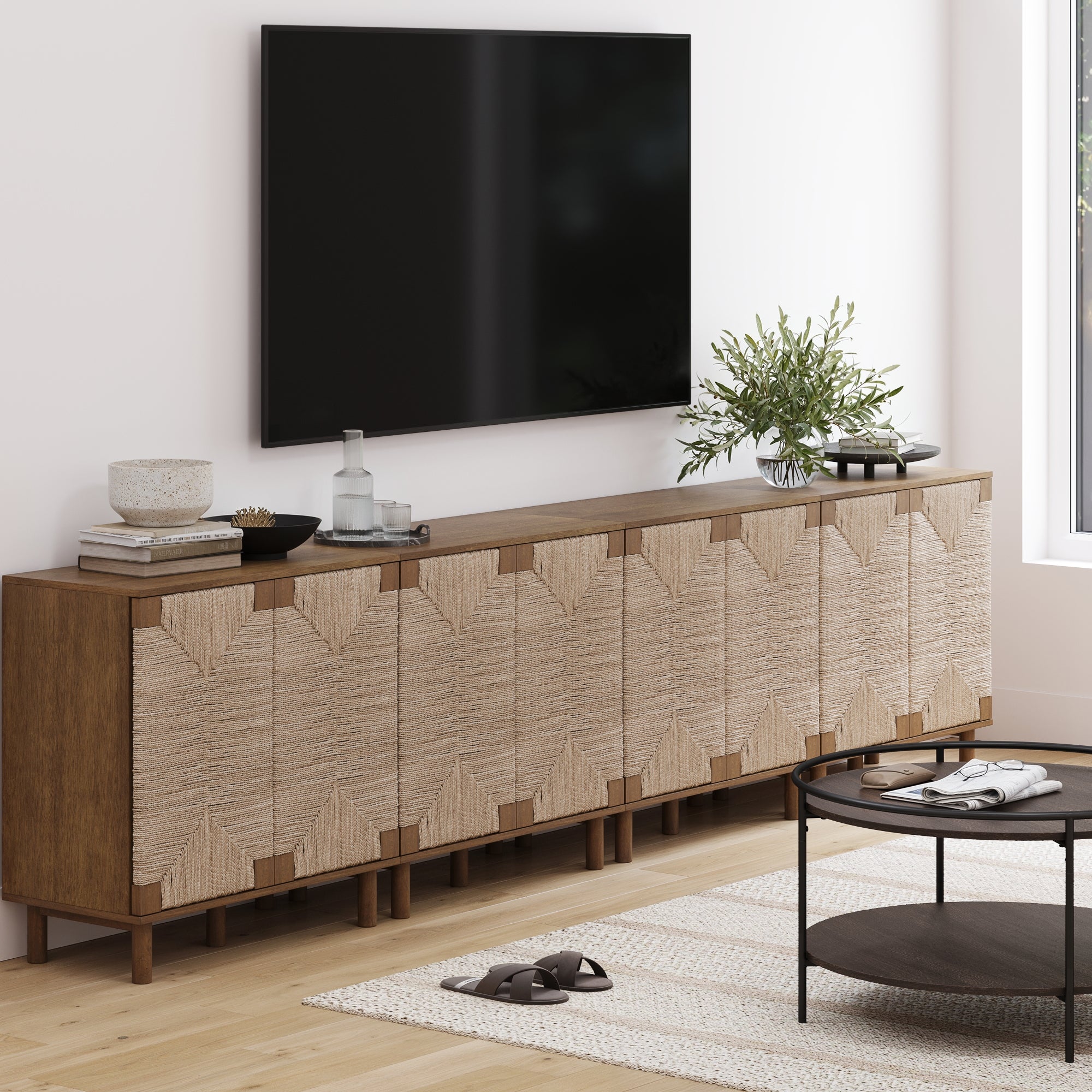 A modern living room features a Large Seagrass TV Console Credenza in dark brown by Nathan James beneath a wall-mounted TV, with decor items on top, a round coffee table, and a woven rug on a light wood floor.
