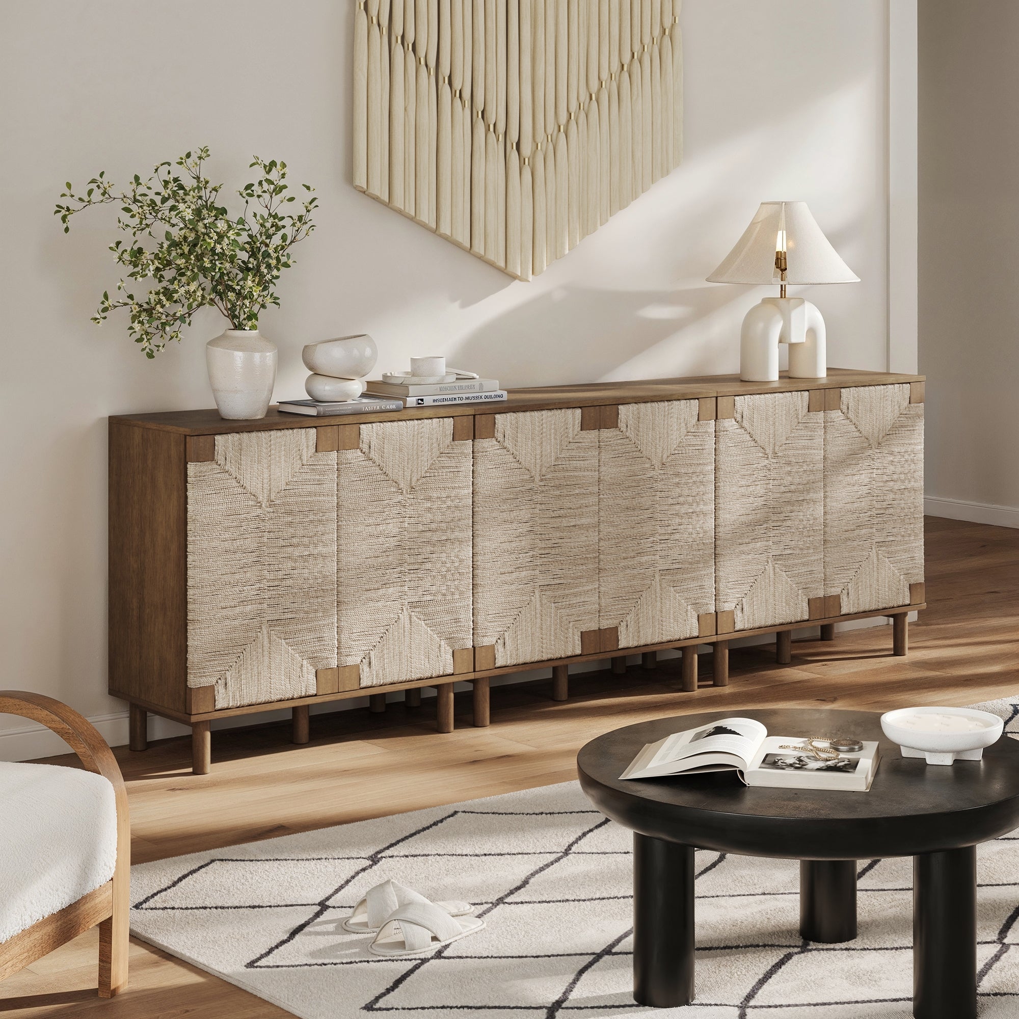A modern living room with a wooden sideboard featuring textured, geometric cabinet fronts, decorated with vases, a lamp, and greenery. A round black coffee table and patterned rug are in the foreground.