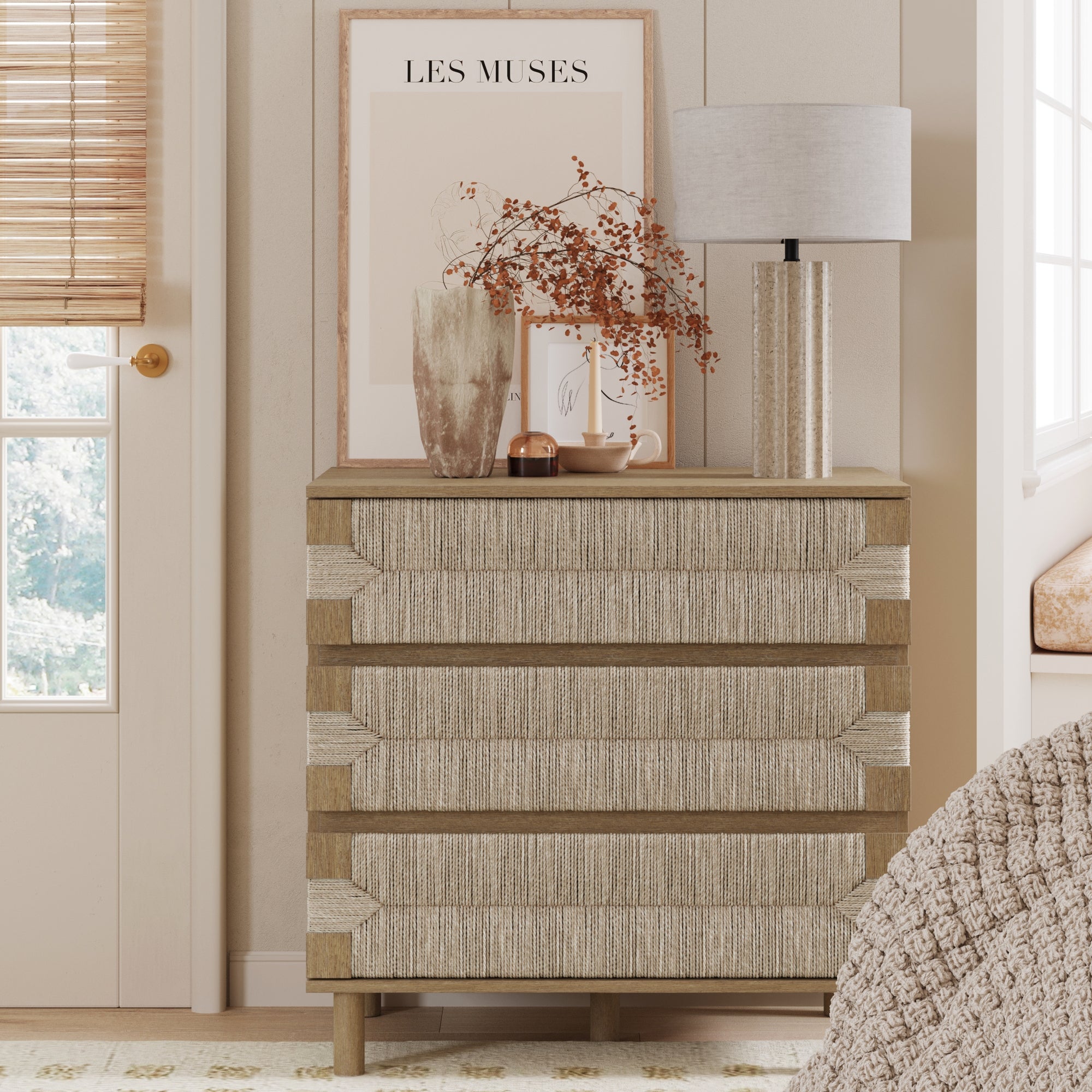 A chest of drawers with a textured front sits against a wall. On top are a lamp, vase with dried flowers, cup, and framed art print reading LES MUSES. Light streams from a nearby window with blinds.