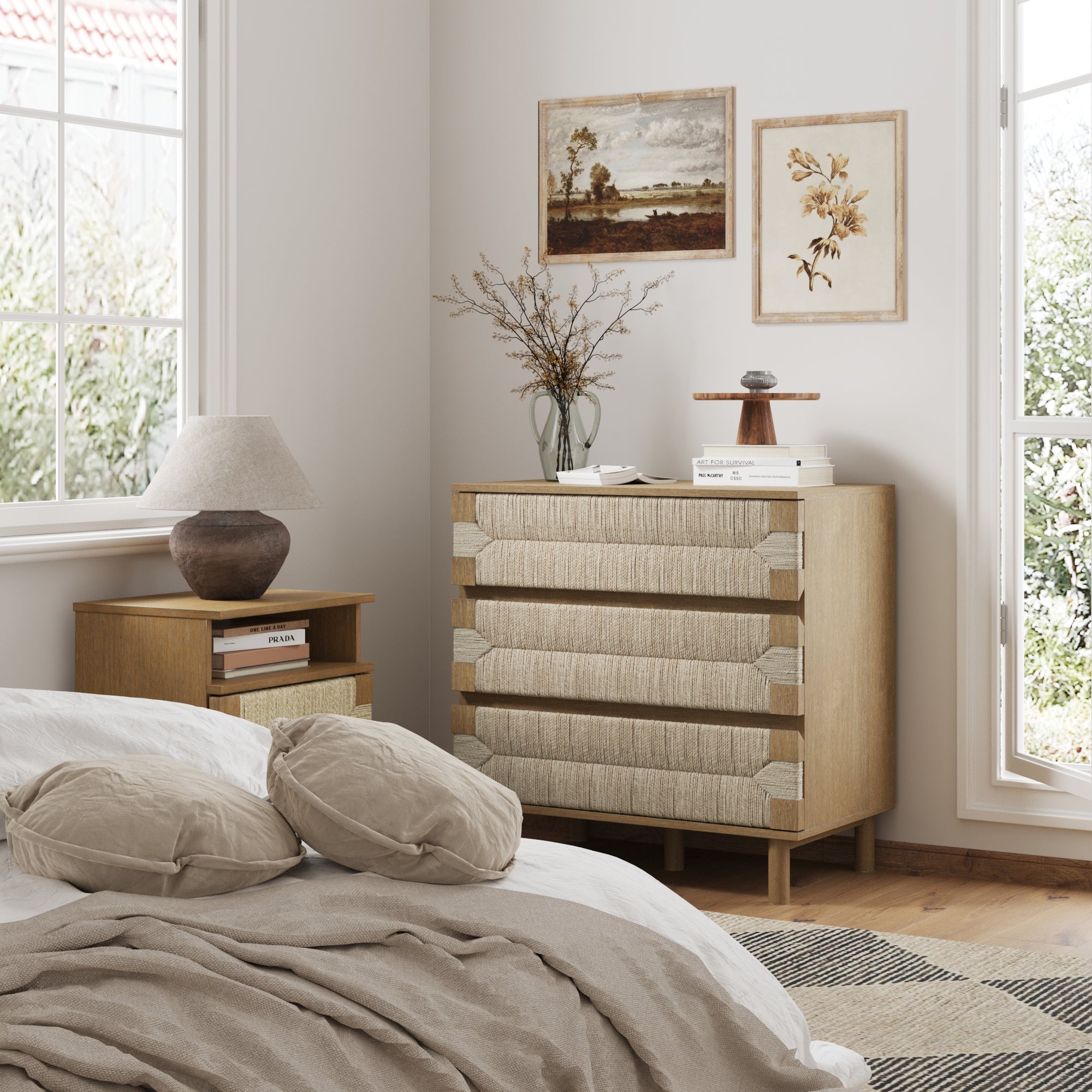 A cozy bedroom corner with a bed, beige cushions, a wooden nightstand with lamp, and the Nathan James Boho Seagrass 3-Drawer Dresser in light brown. Sunlight streams in on two framed artworks displayed on the wall.